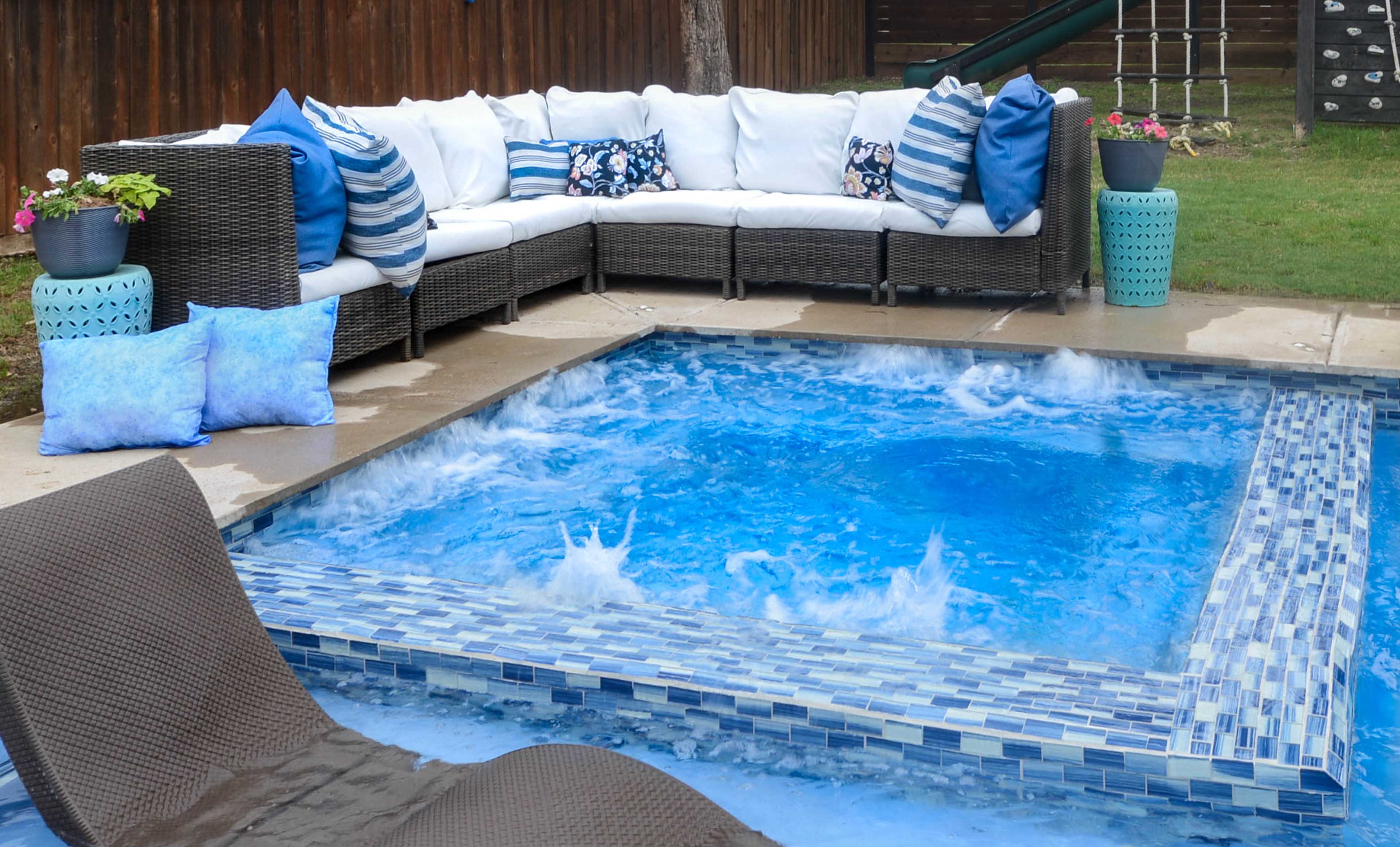 The image shows a backyard setting featuring a rectangular pool with blue tiled edges, surrounded by a sectional wicker sofa with decorative pillows and a few lounge chairs nearby.