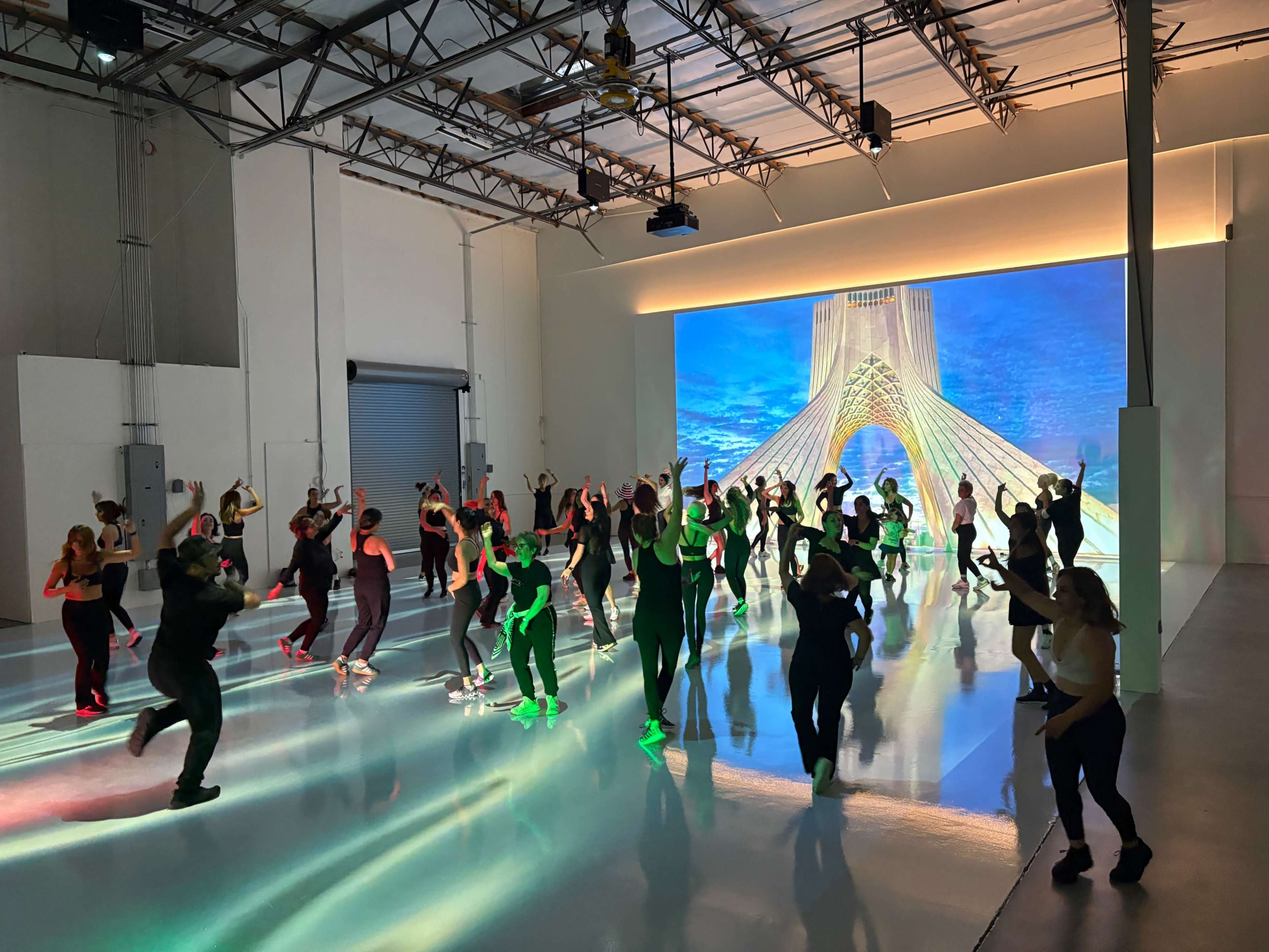 A group of individuals is participating in a lively dance class in a spacious studio with bright lights and a large projection of a bridge in the background.
