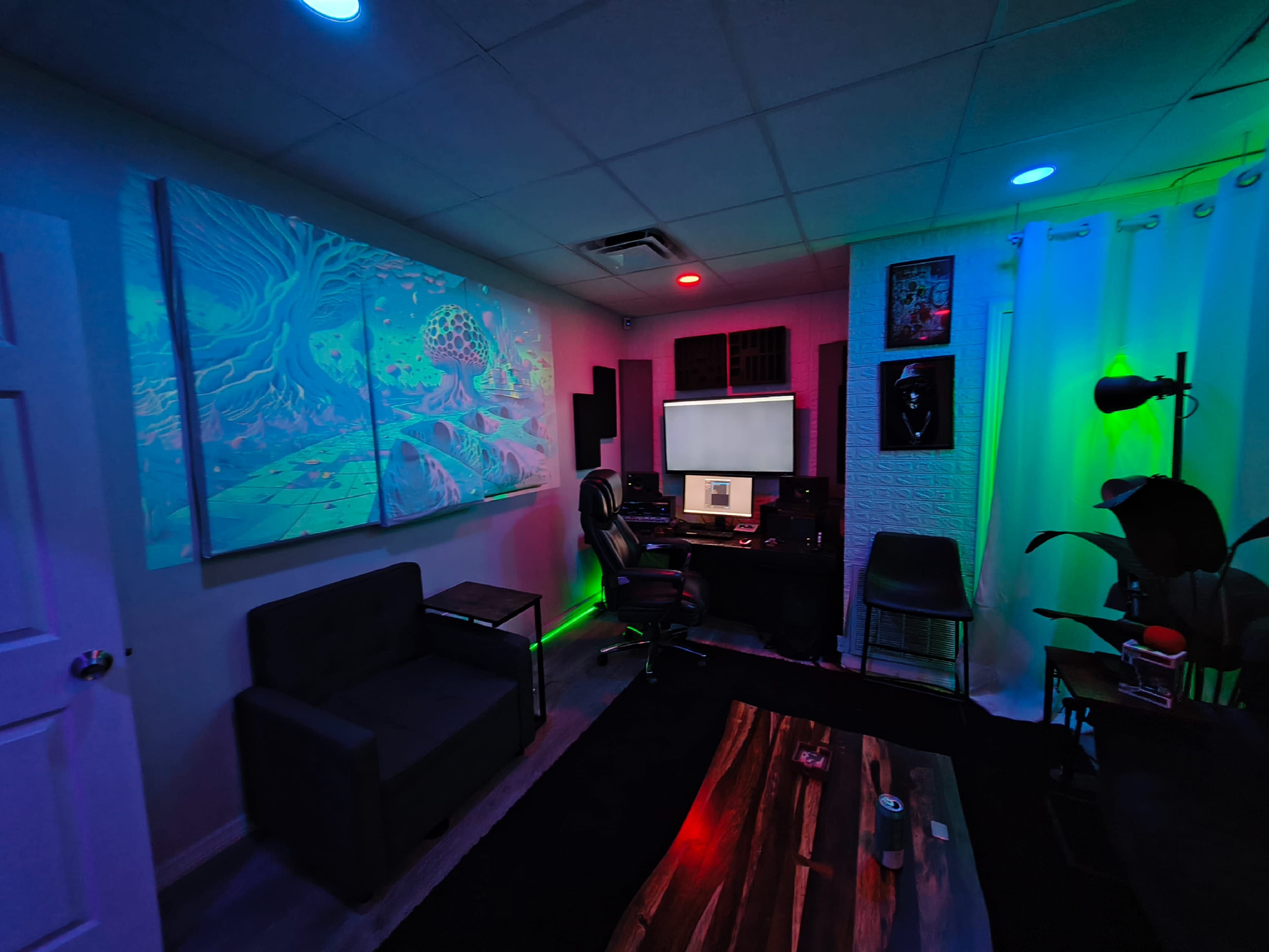 The image shows a dimly lit room featuring a computer desk with monitors, a couch, colorful LED lights, and a large abstract artwork on the wall.