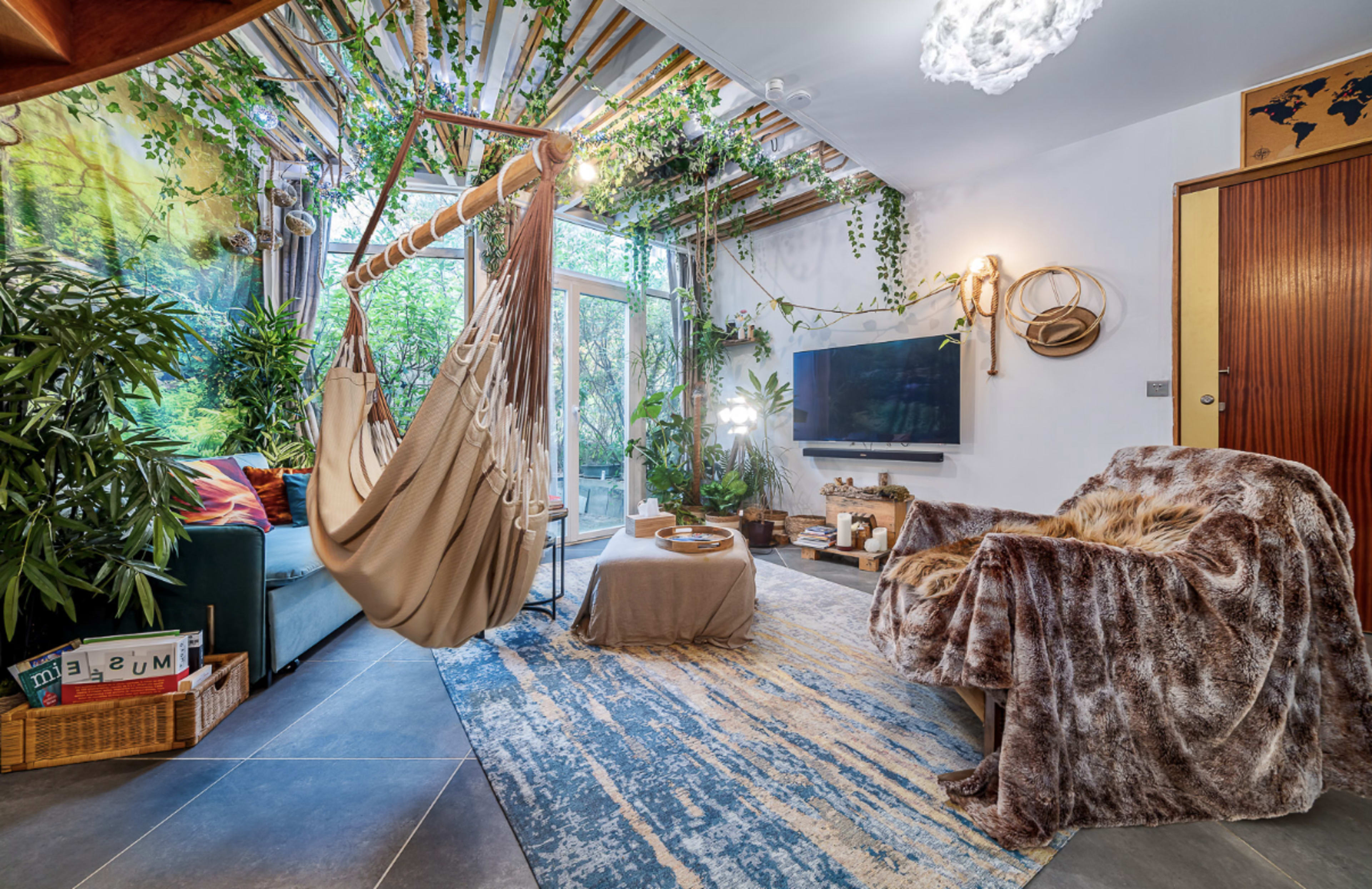 The image shows a cozy living room with a hammock, a large plush chair, green plants, and a TV against a backdrop of a nature-themed wall.