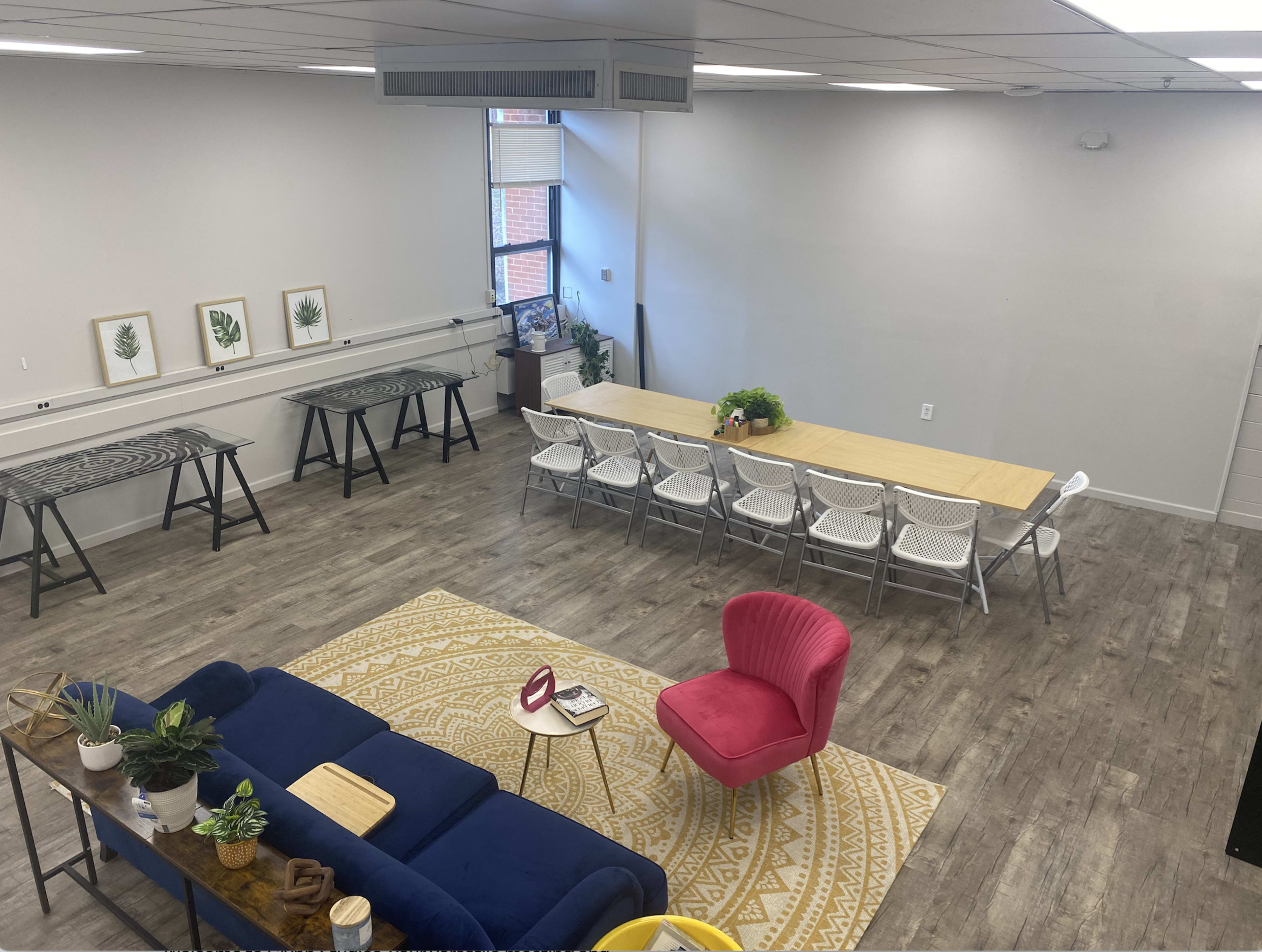 A spacious, modern communal area featuring a long dining table surrounded by chairs, a seating area with a blue sofa and a pink armchair, and several worktables against the walls.