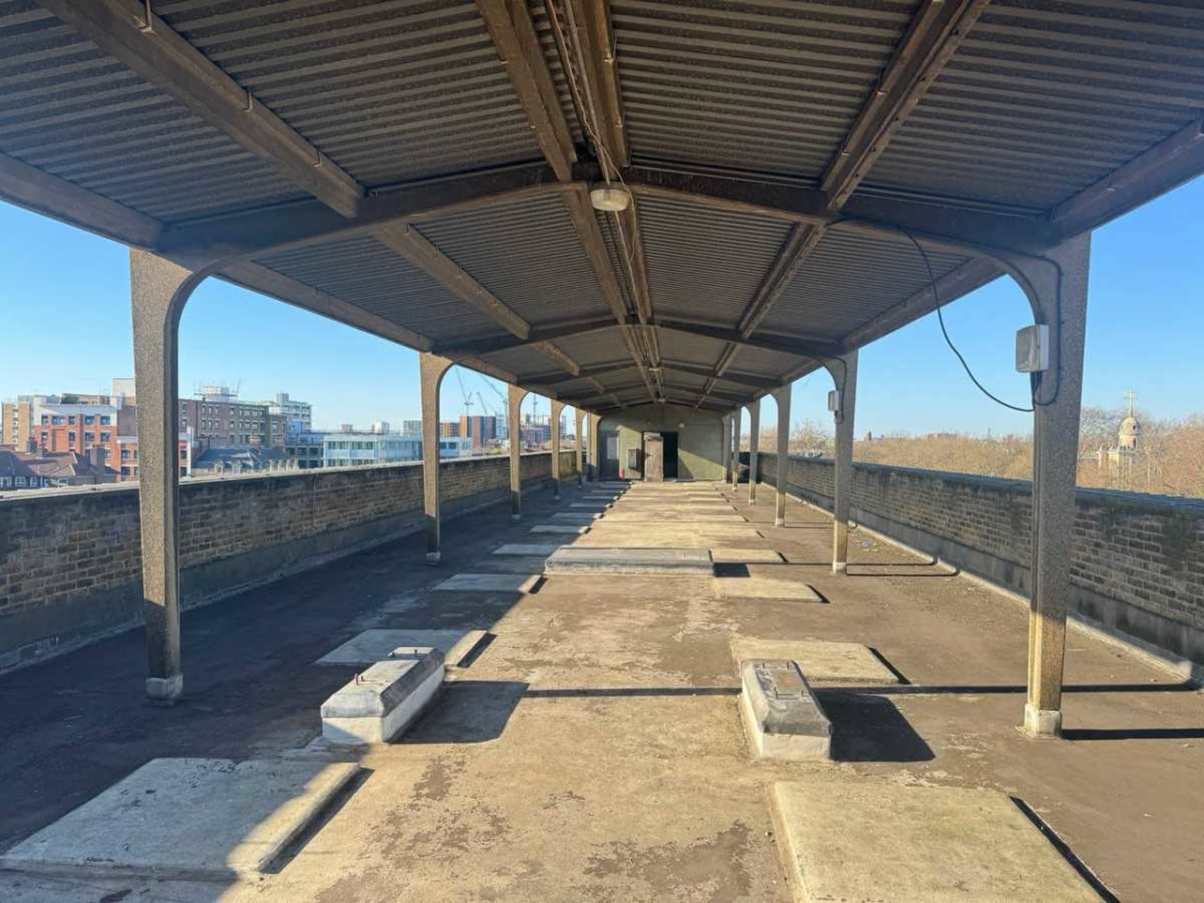 The image shows a long, empty walkway with a metal roof supported by columns, overlooking a cityscape in the background.
