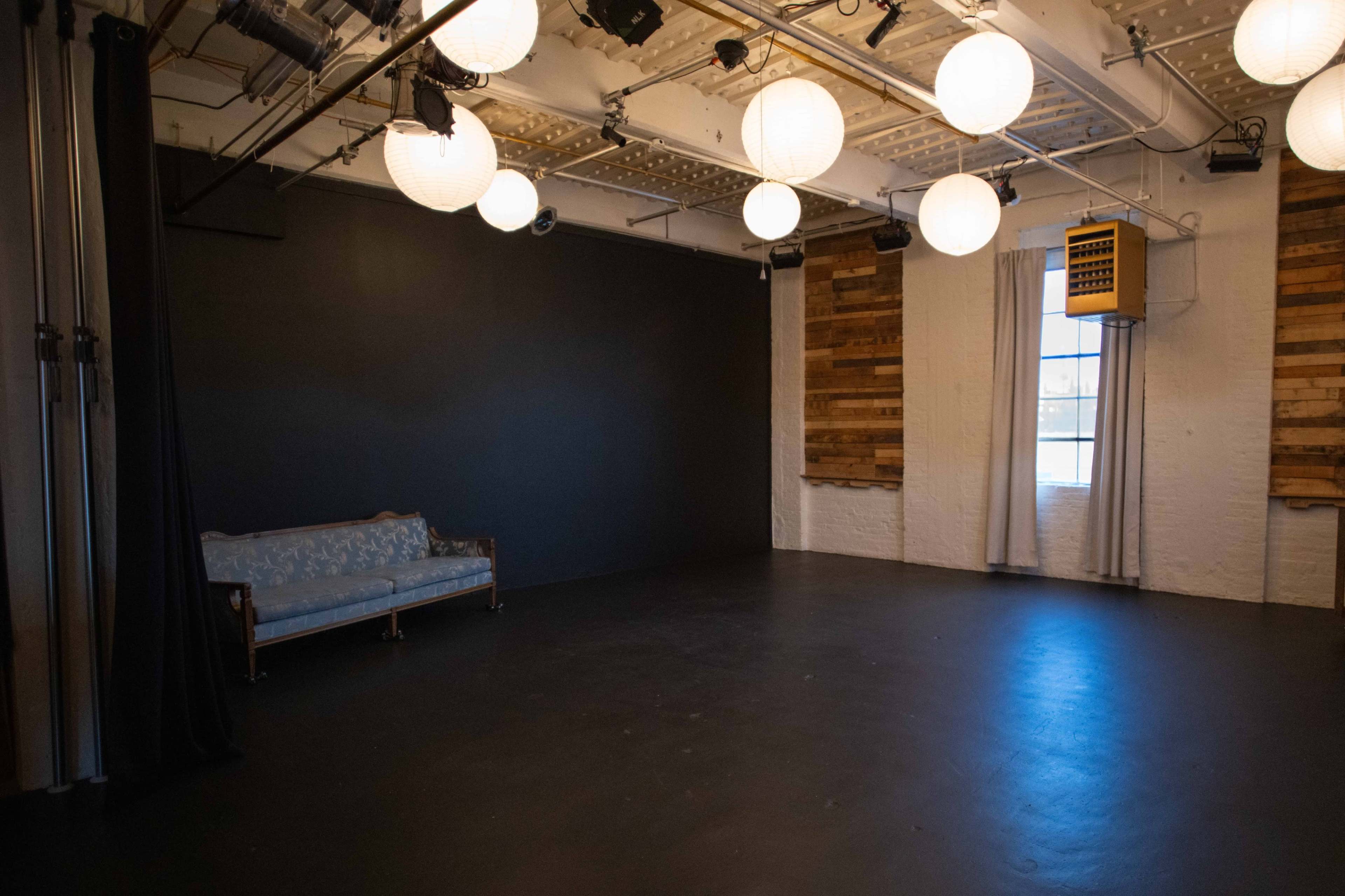 The image shows a spacious room with a gray floor, a wooden couch against one wall, and round pendant lights overhead.