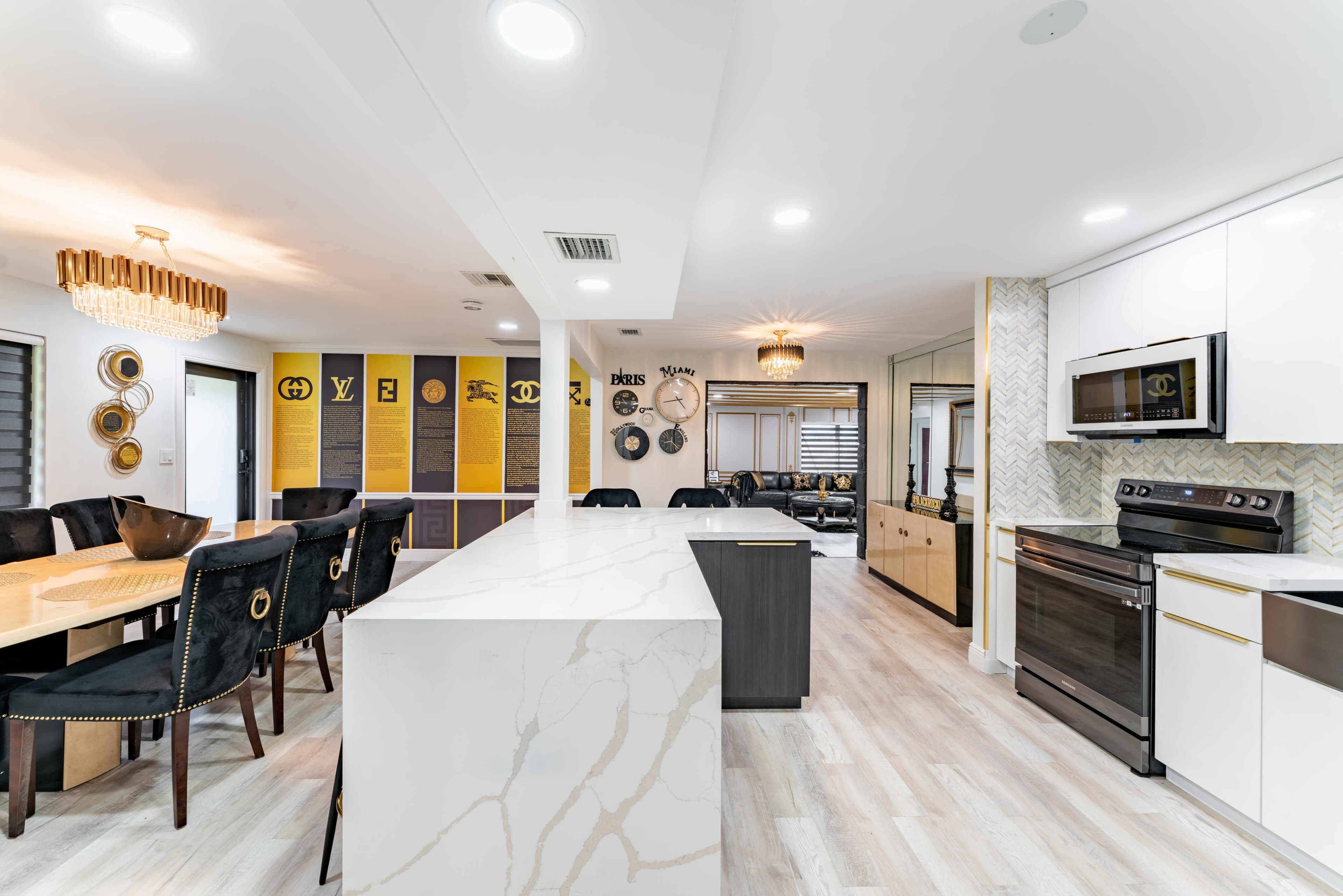 The image shows a modern kitchen and dining area with a large marble island, stainless steel appliances, and a dark dining table surrounded by upholstered chairs.