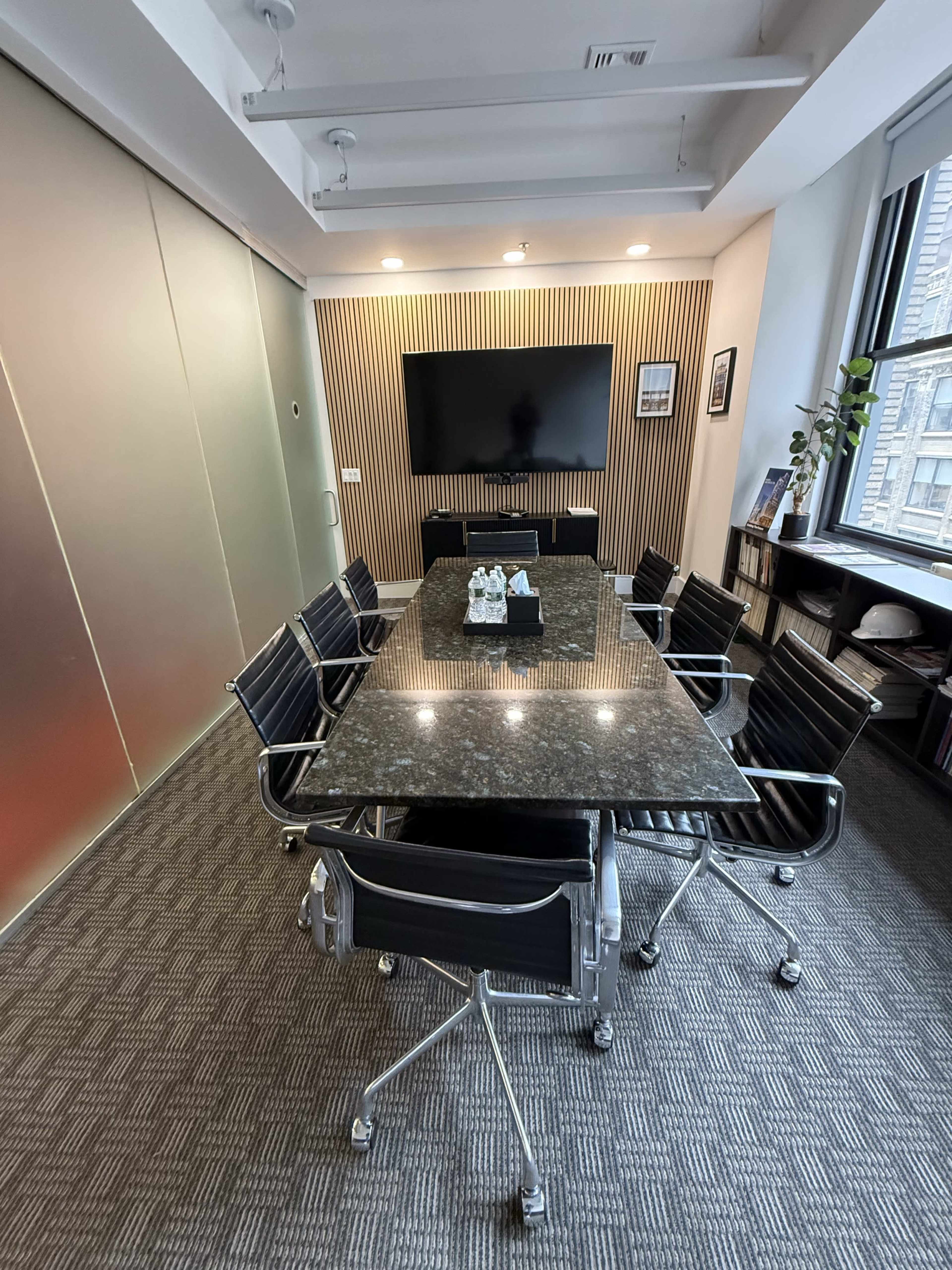 The image shows a modern conference room with a long table surrounded by black chairs, a television screen mounted on the wall, and large windows offering a view of the outside.