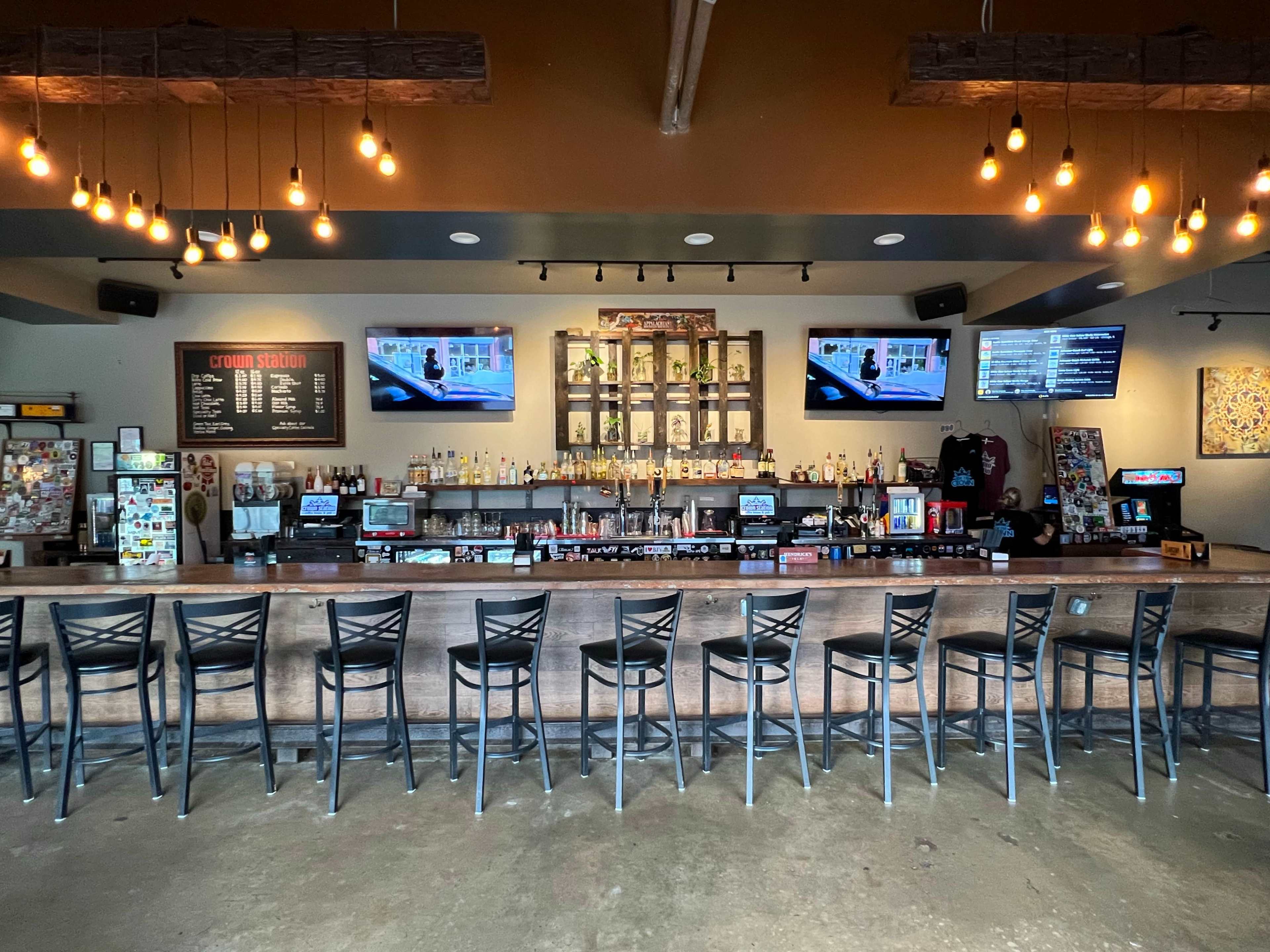 A well-lit bar with a long wooden counter, multiple rows of bar stools, and shelves stocked with liquor, flanked by two televisions displaying content.