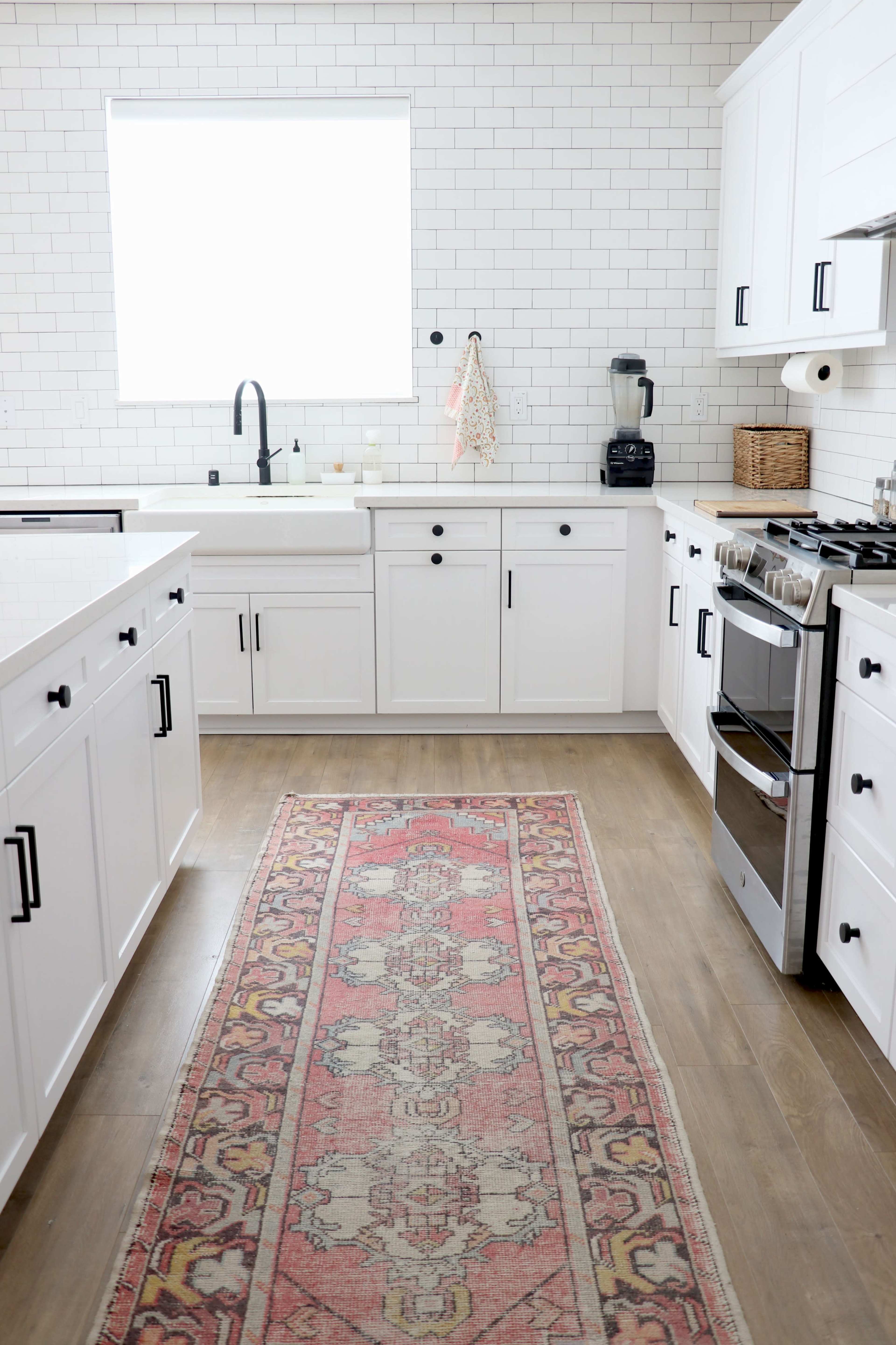 A contemporary kitchen features white cabinets, a large window, and a patterned rug on the hardwood floor.