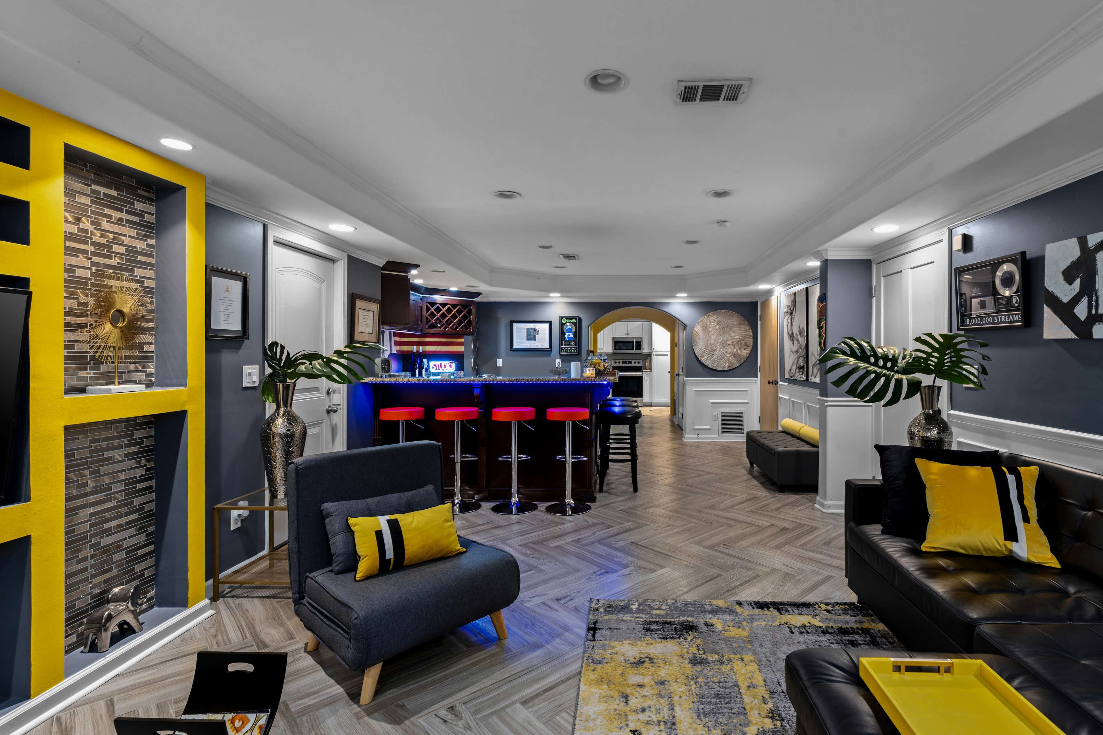 The image shows a modern basement lounge with a wet bar, comfortable seating, and decorative accents in yellow and gray tones.