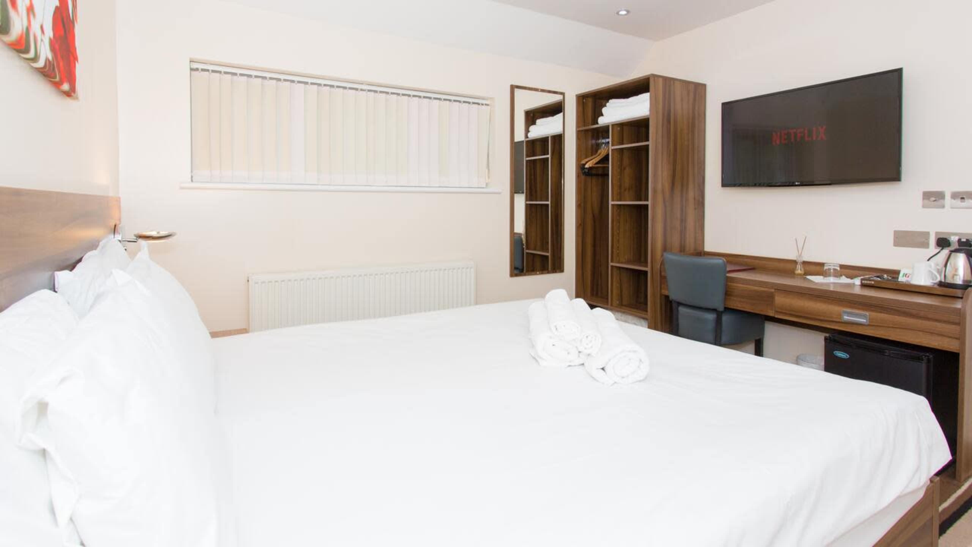 A simple hotel room with a bed, a small desk, a television displaying the Netflix logo, and a wardrobe.