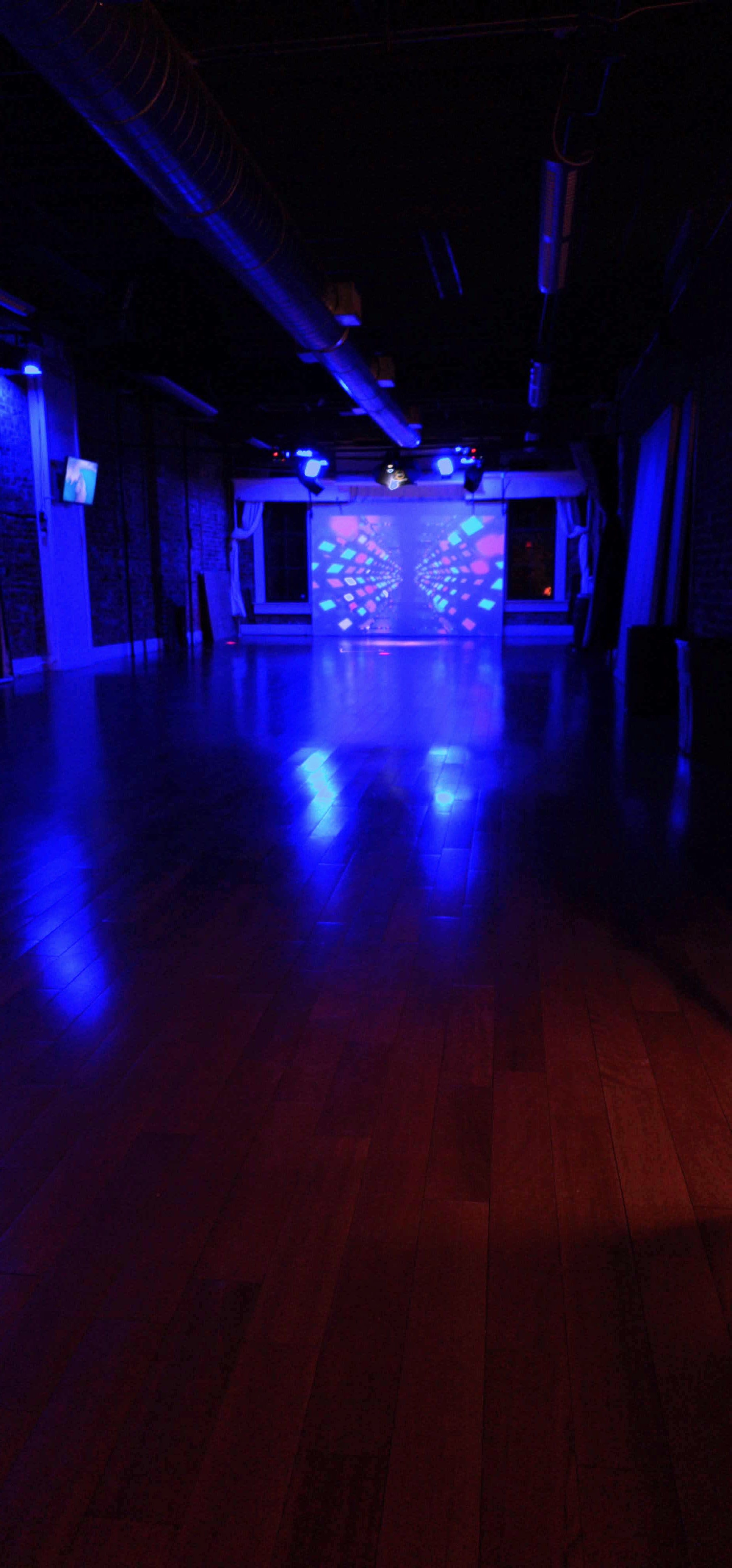 The image shows an empty dance studio with a polished wooden floor, illuminated by blue and colorful lights reflecting off a wall.