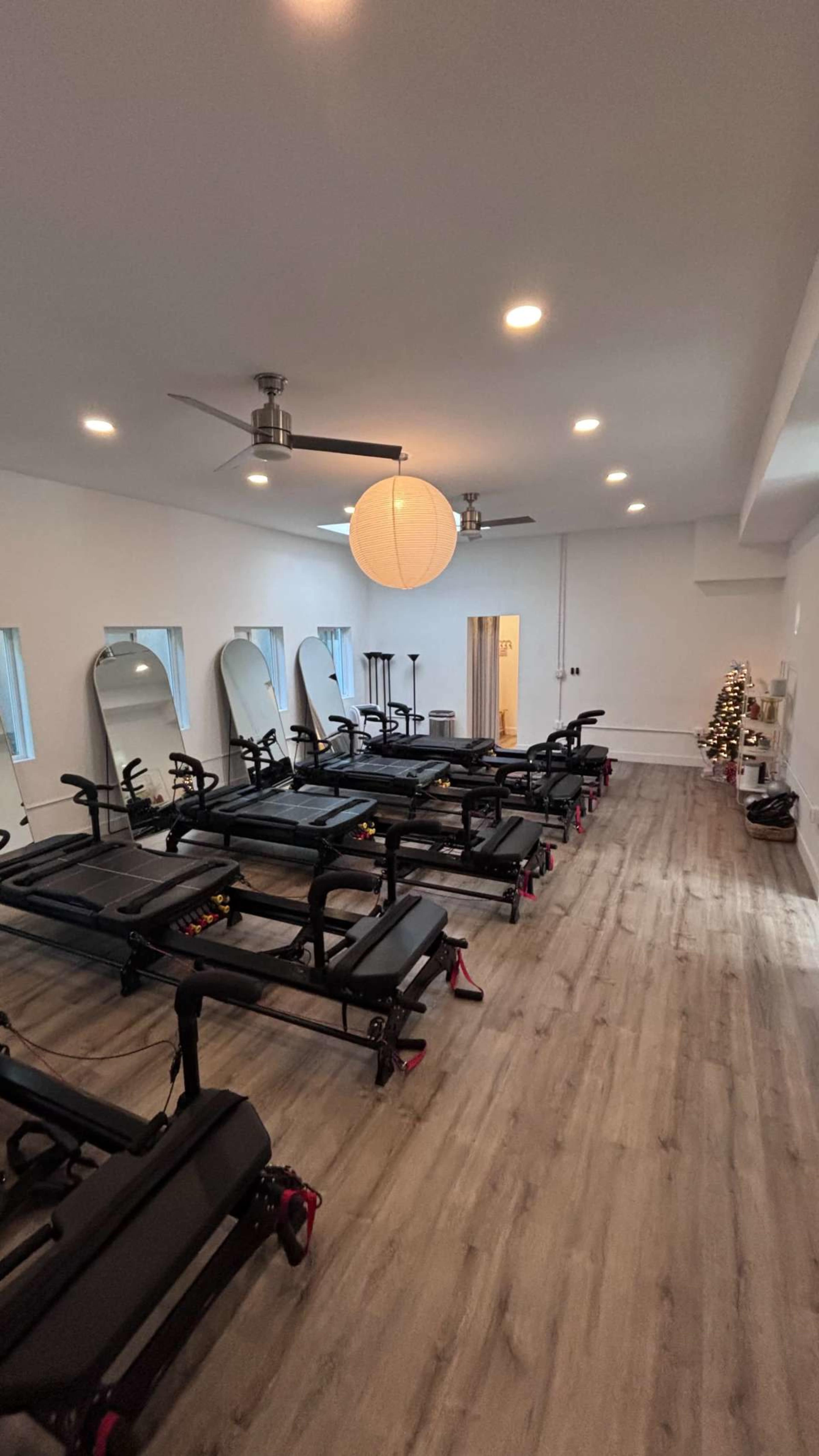 The image shows an empty fitness studio equipped with several reformer machines arranged in rows, with mirrors and decorative lighting.