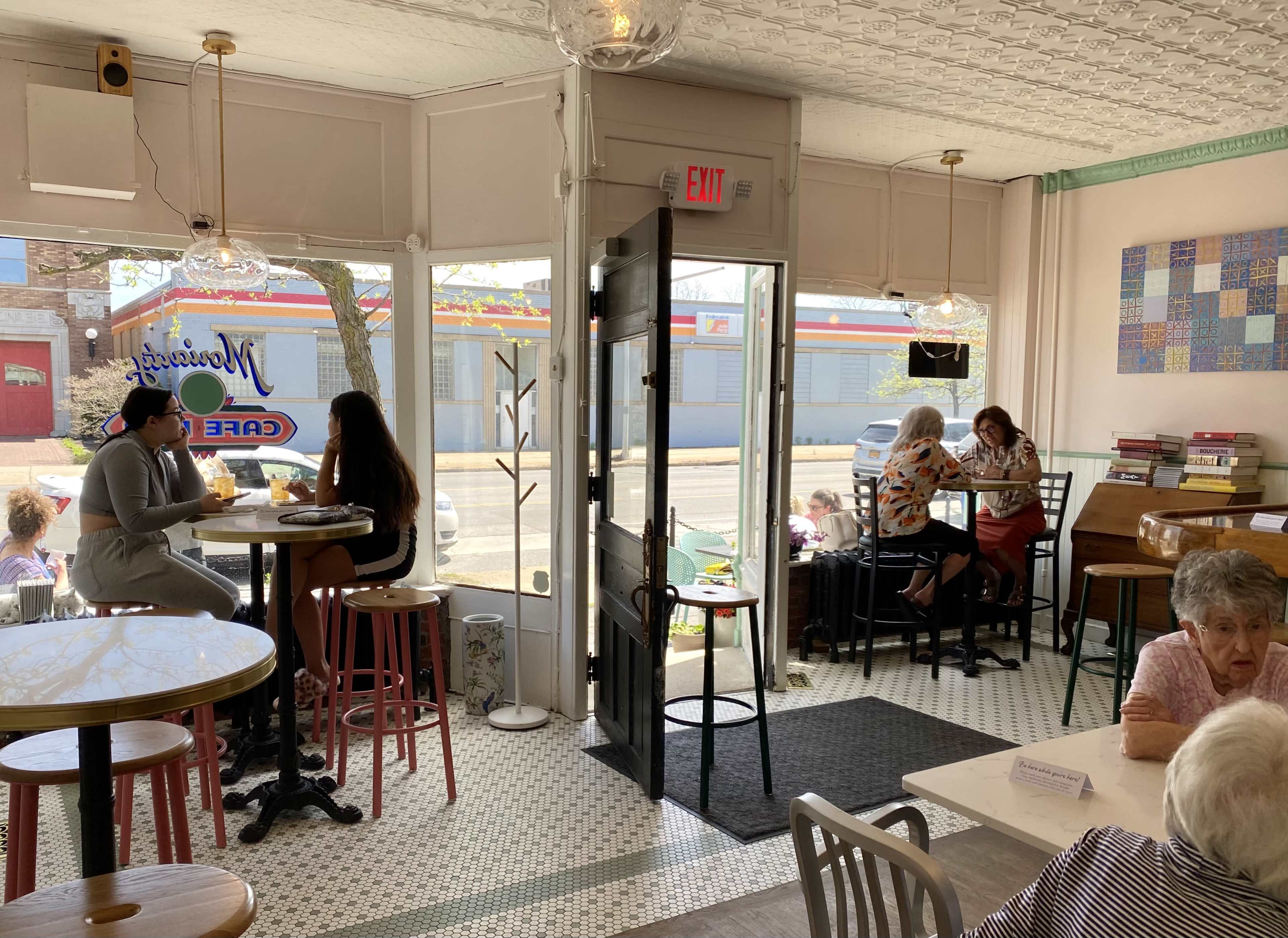 A café interior with various seating arrangements, including two women at a table by the window and a group of patrons in the background.