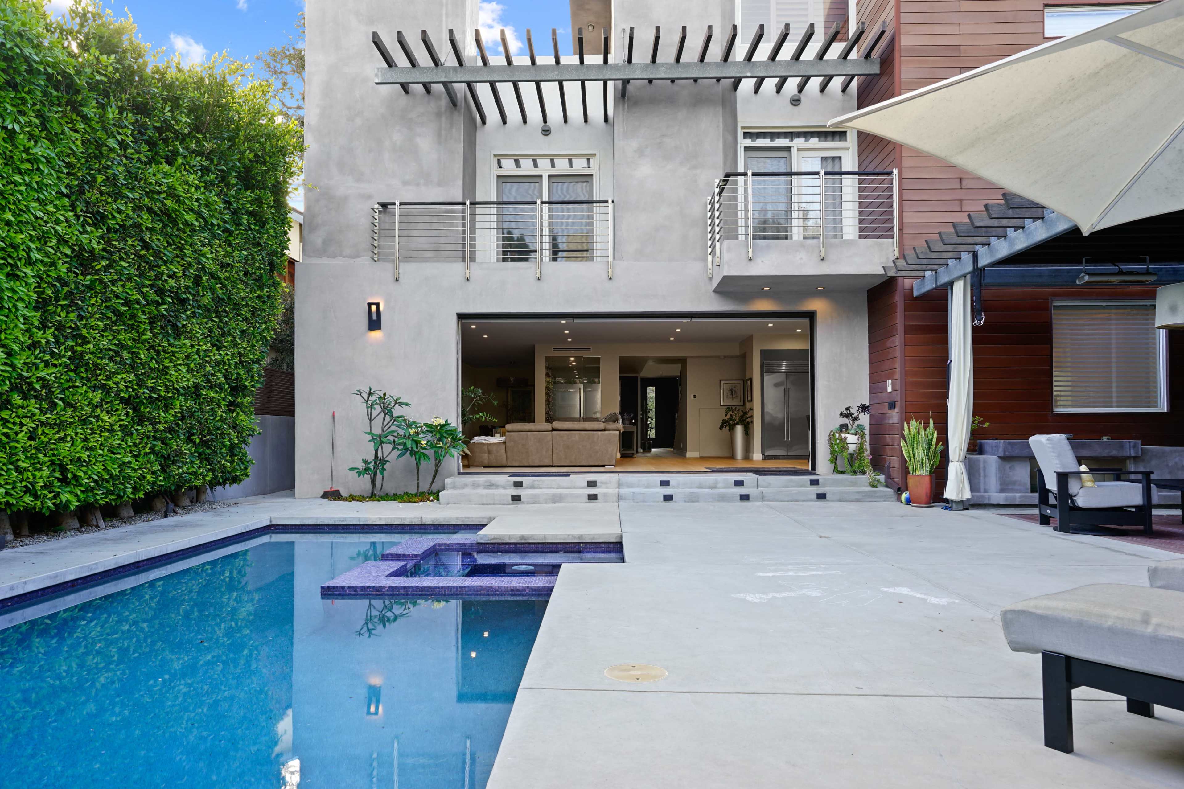 The image shows a modern house with a pool area surrounded by concrete and greenery, featuring a large living space visible through sliding glass doors.