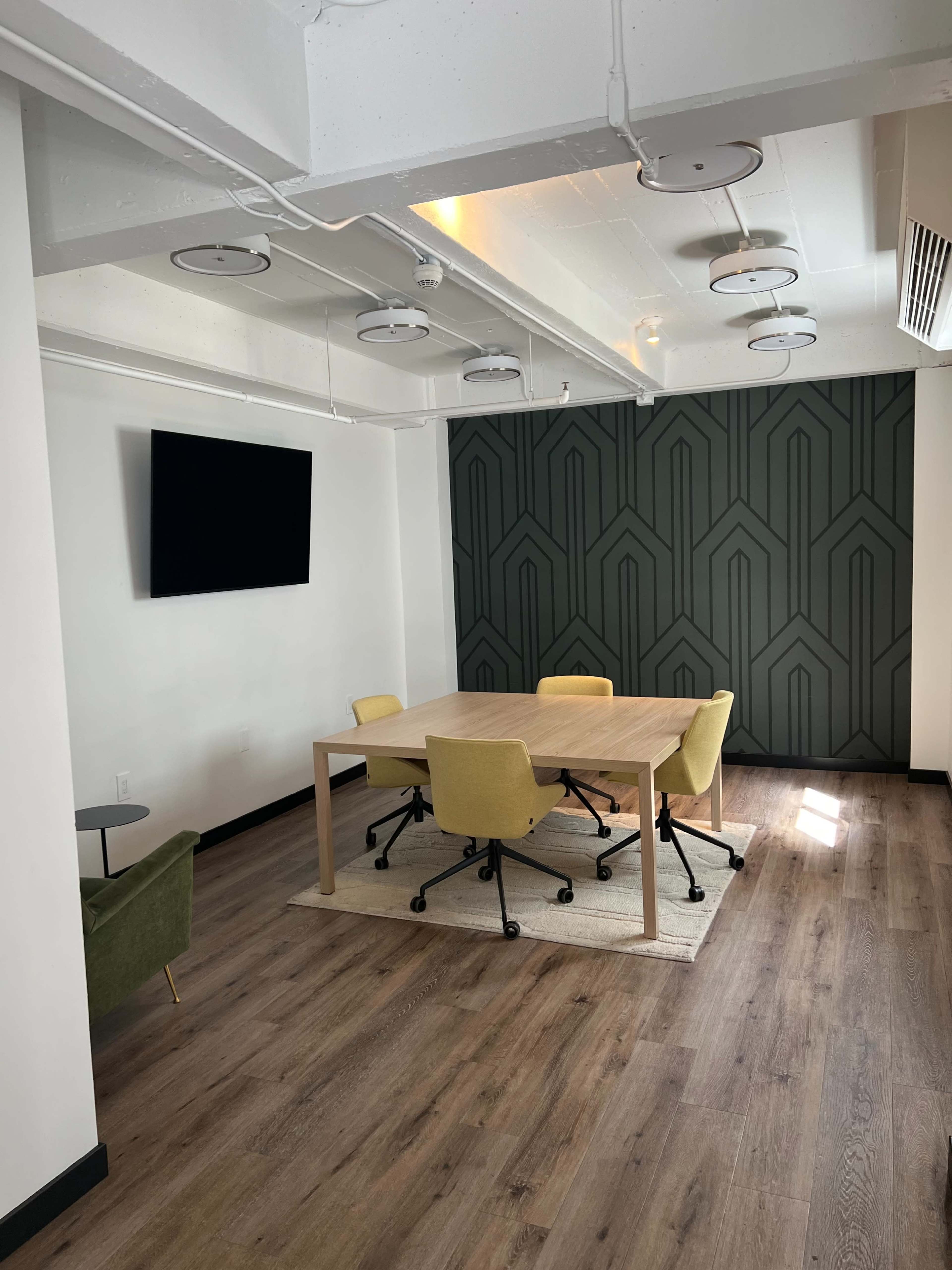 A modern meeting room features a wooden table surrounded by yellow chairs, with a large TV mounted on a wall and a textured green accent wall in the background.