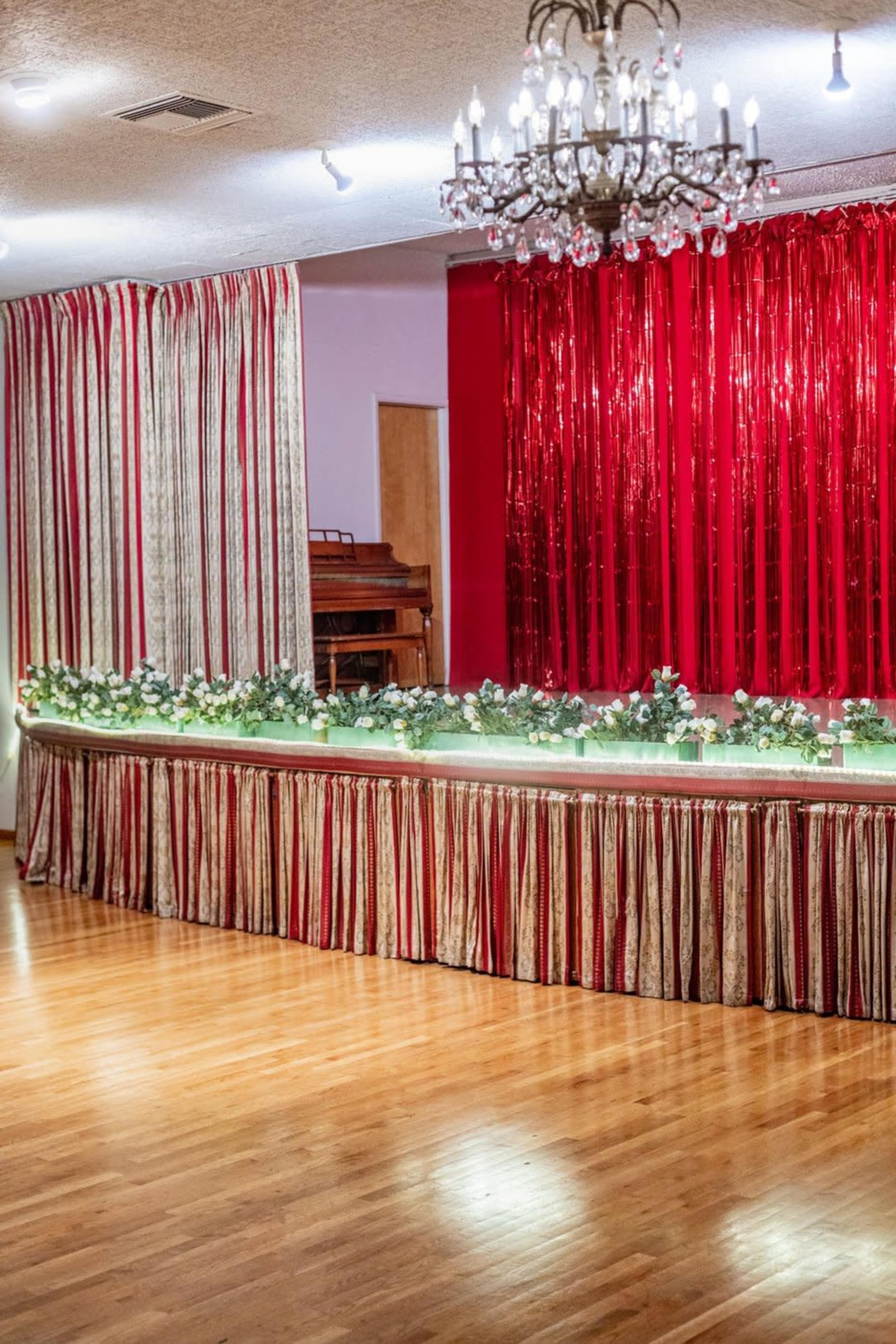 The image shows a decorated stage area with a red and gold curtain backdrop, a floral arrangement along a table, and a chandelier overhead.
