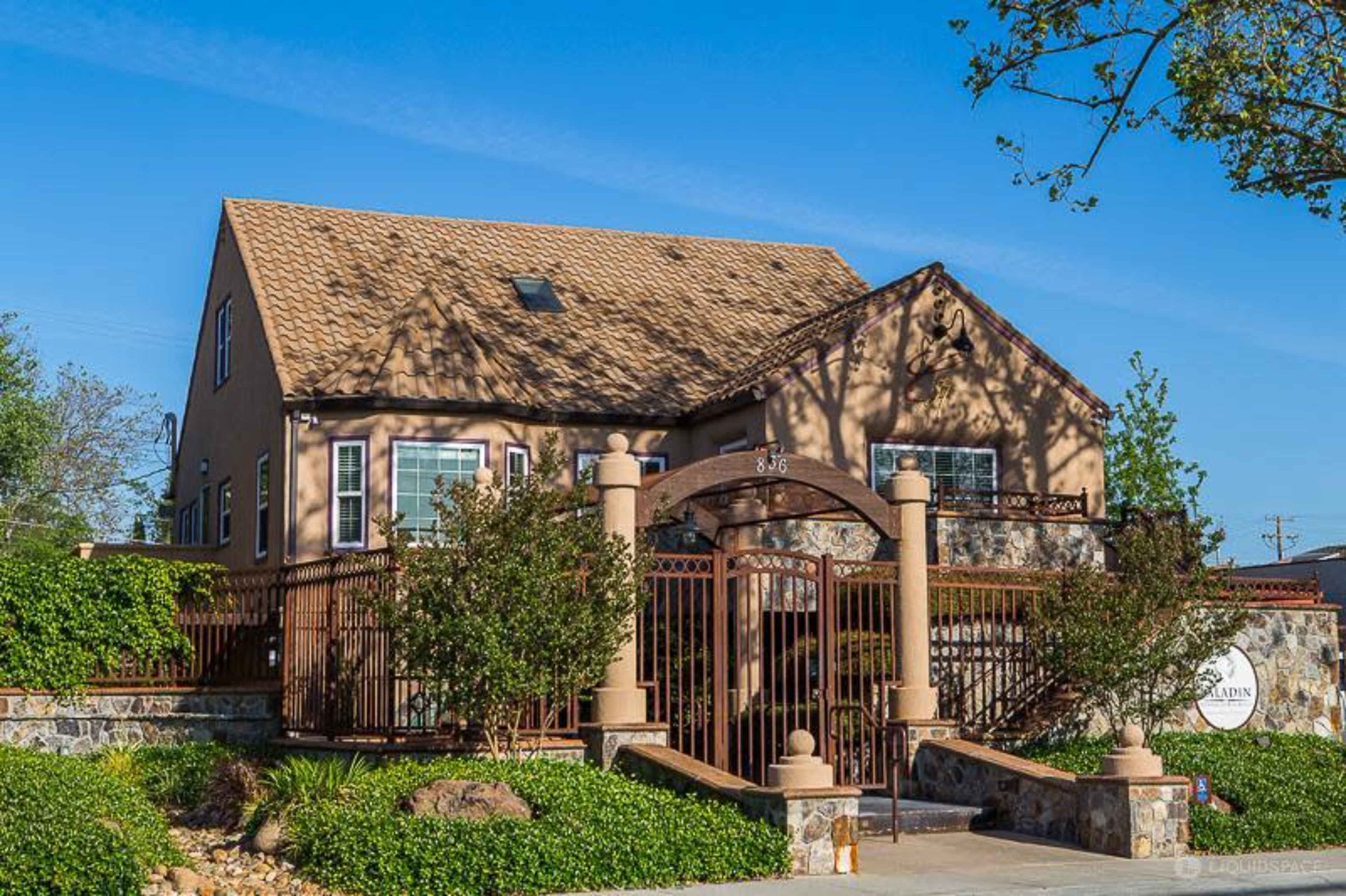 A two-story house with a brown tiled roof, surrounded by green landscaping and a decorative metal fence.