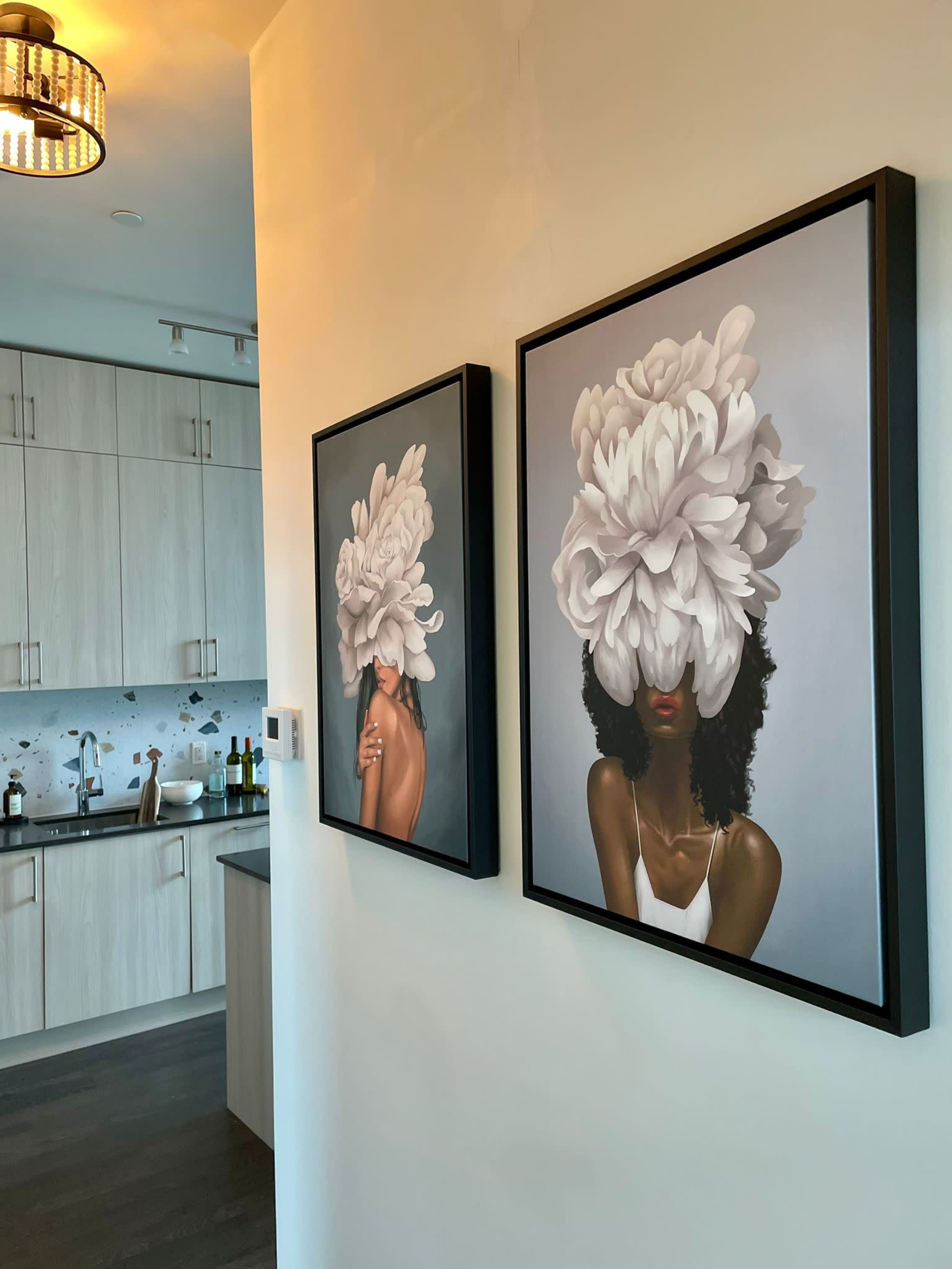 Two framed artworks depicting women with large floral arrangements covering their heads are displayed on a wall next to a modern kitchen.