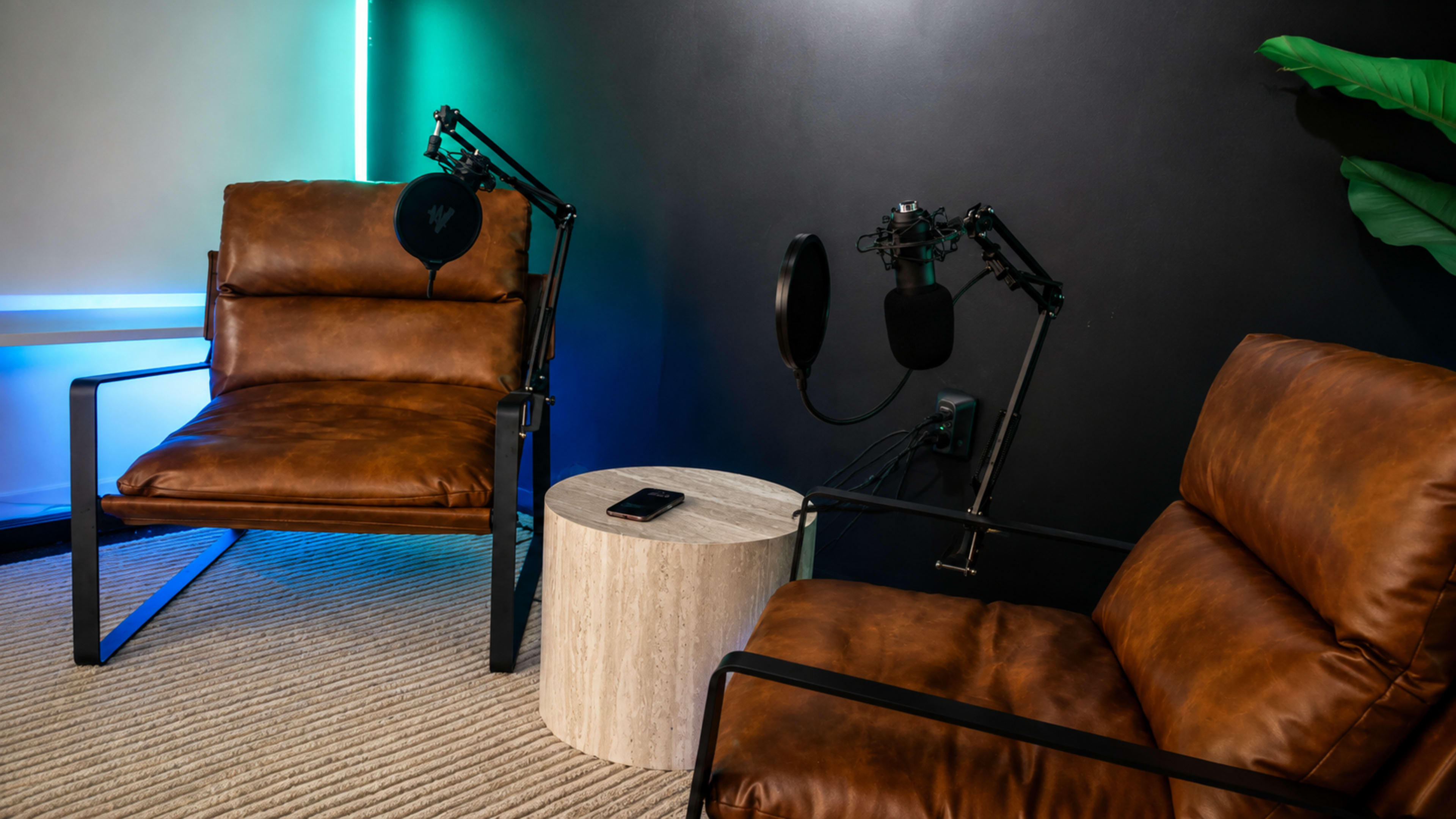 The image features two brown leather chairs positioned next to a circular wooden table, with microphones mounted nearby against a dark wall.
