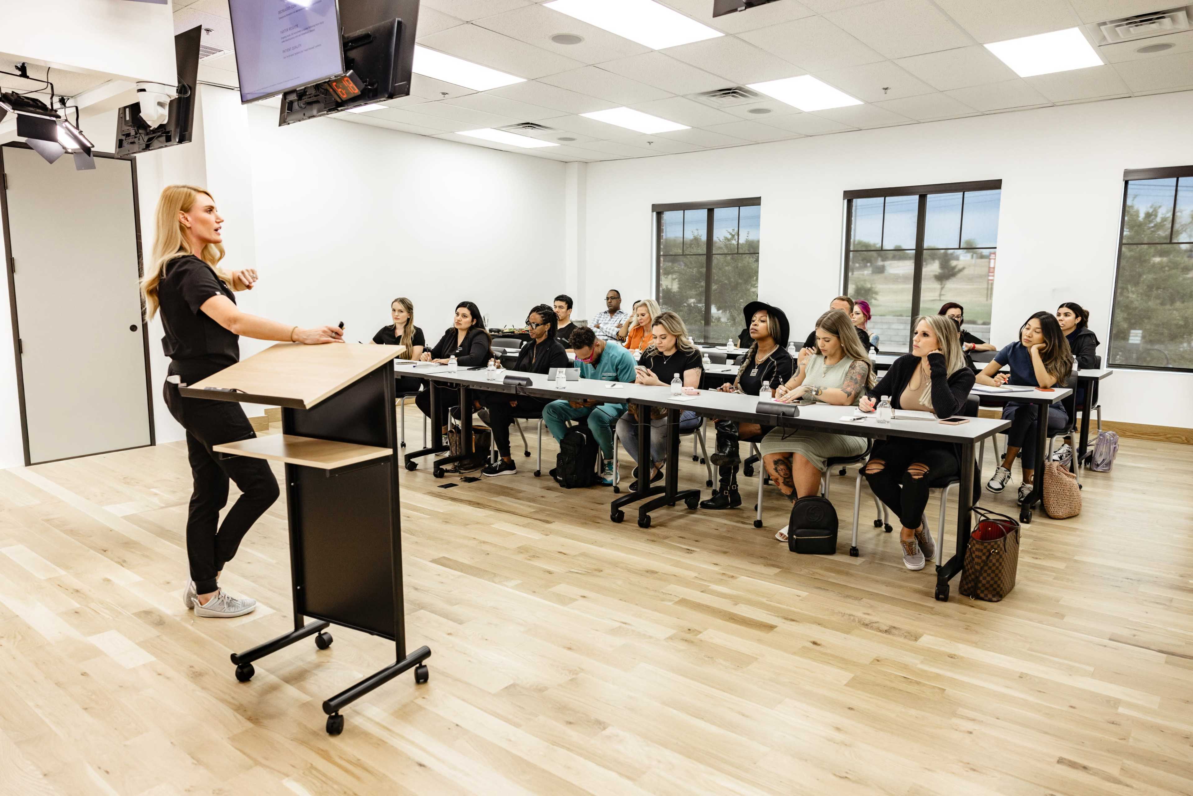 A teacher stands at a podium addressing a class of students seated at desks in a modern classroom.