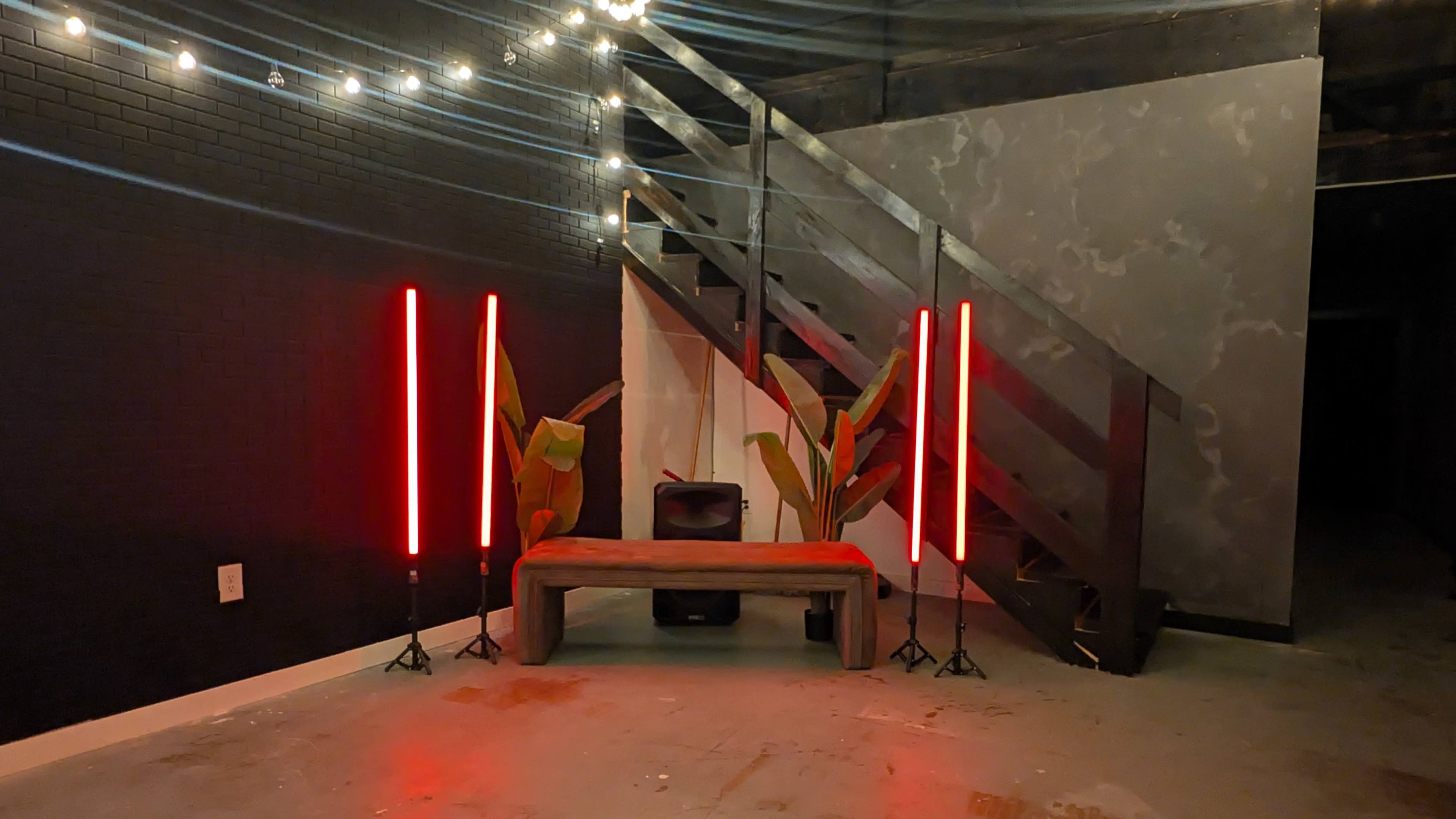 The image shows a dimly lit indoor space featuring a wooden bench, two red vertical light fixtures on stands, and a pair of green plants, with a staircase in the background.