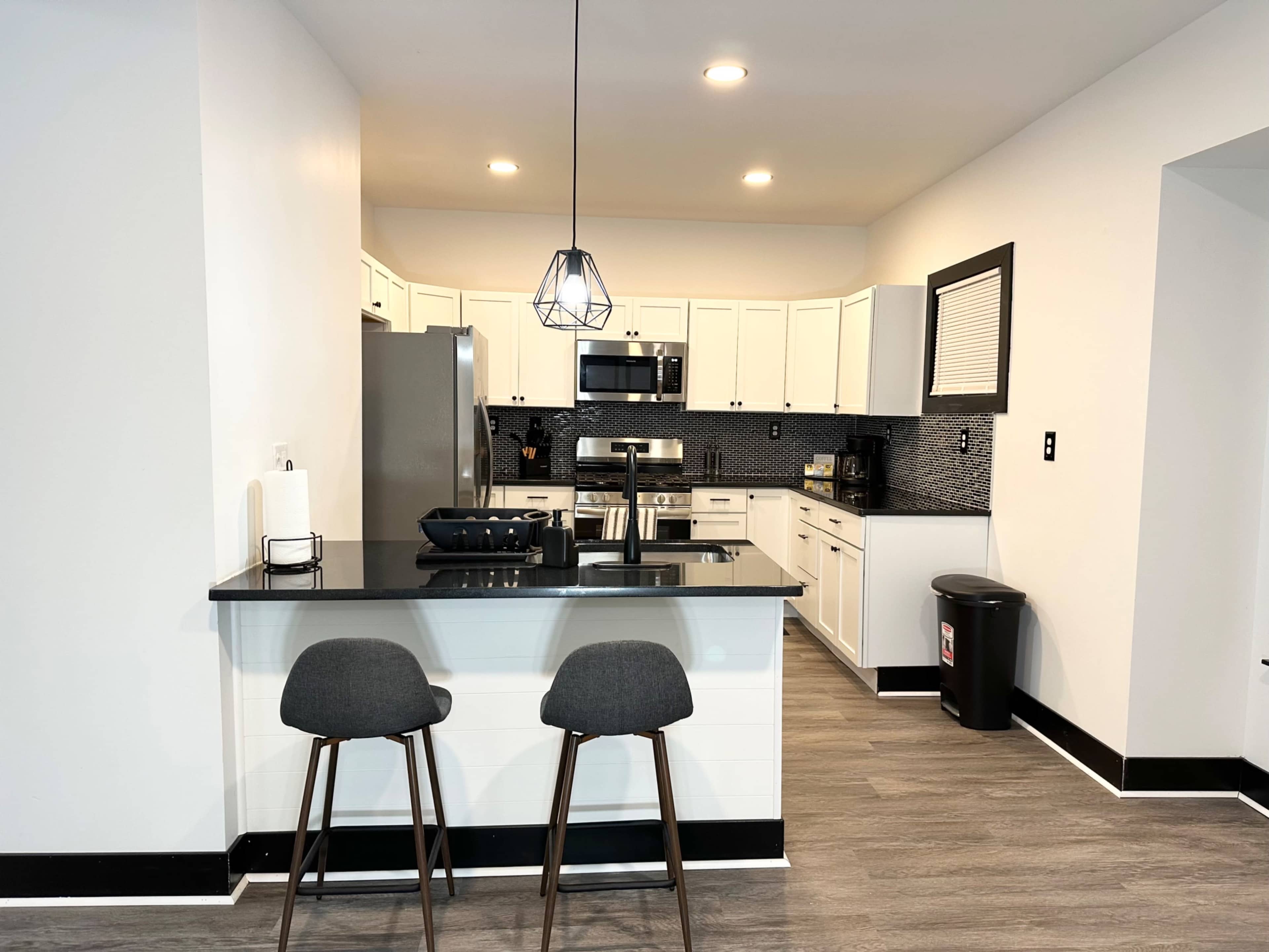 The image shows a modern kitchen featuring white cabinets, stainless steel appliances, and a black countertop with two bar stools.