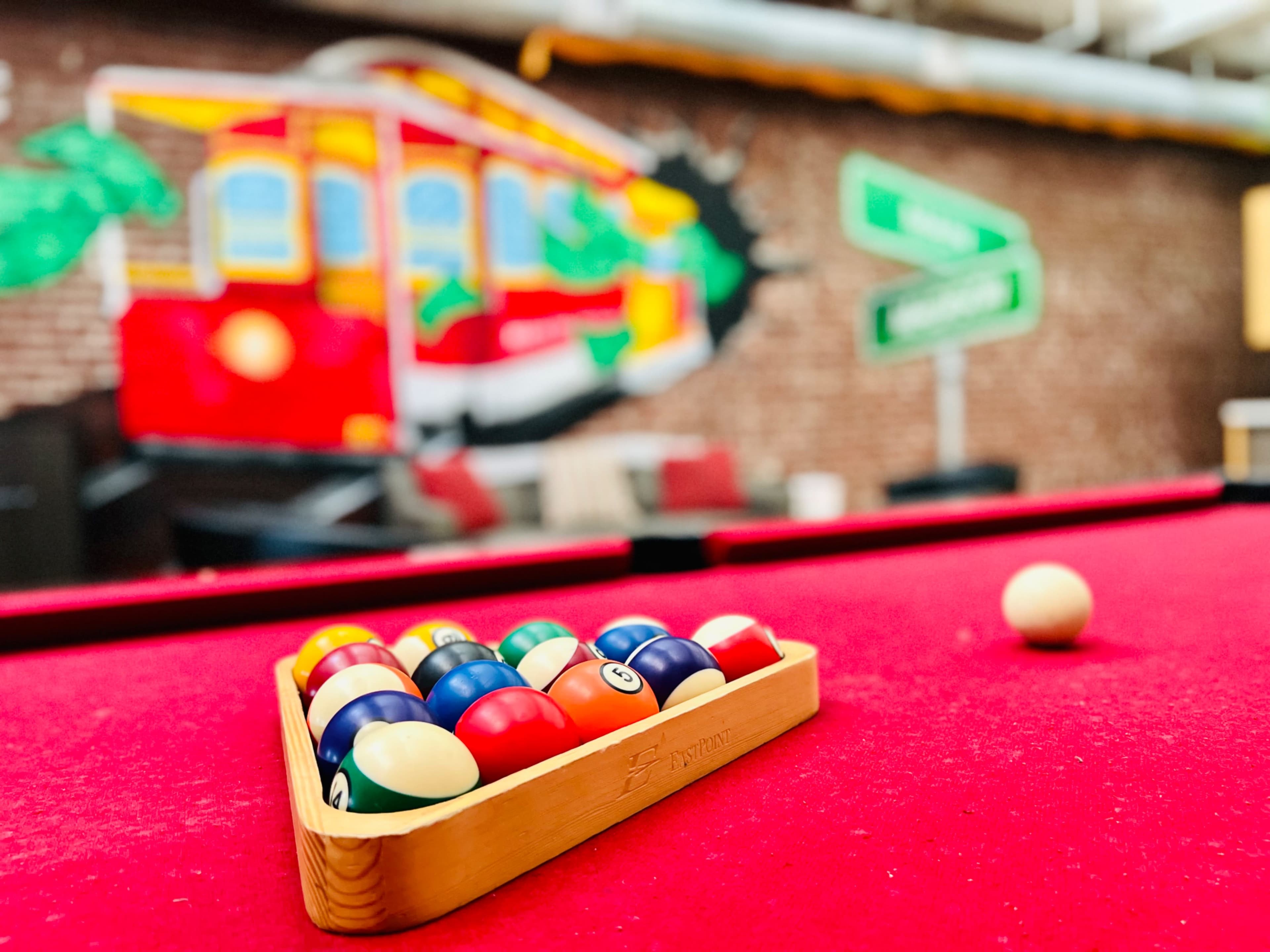 A triangle of billiard balls rests on a red felt pool table in front of a vibrant mural featuring a red streetcar.