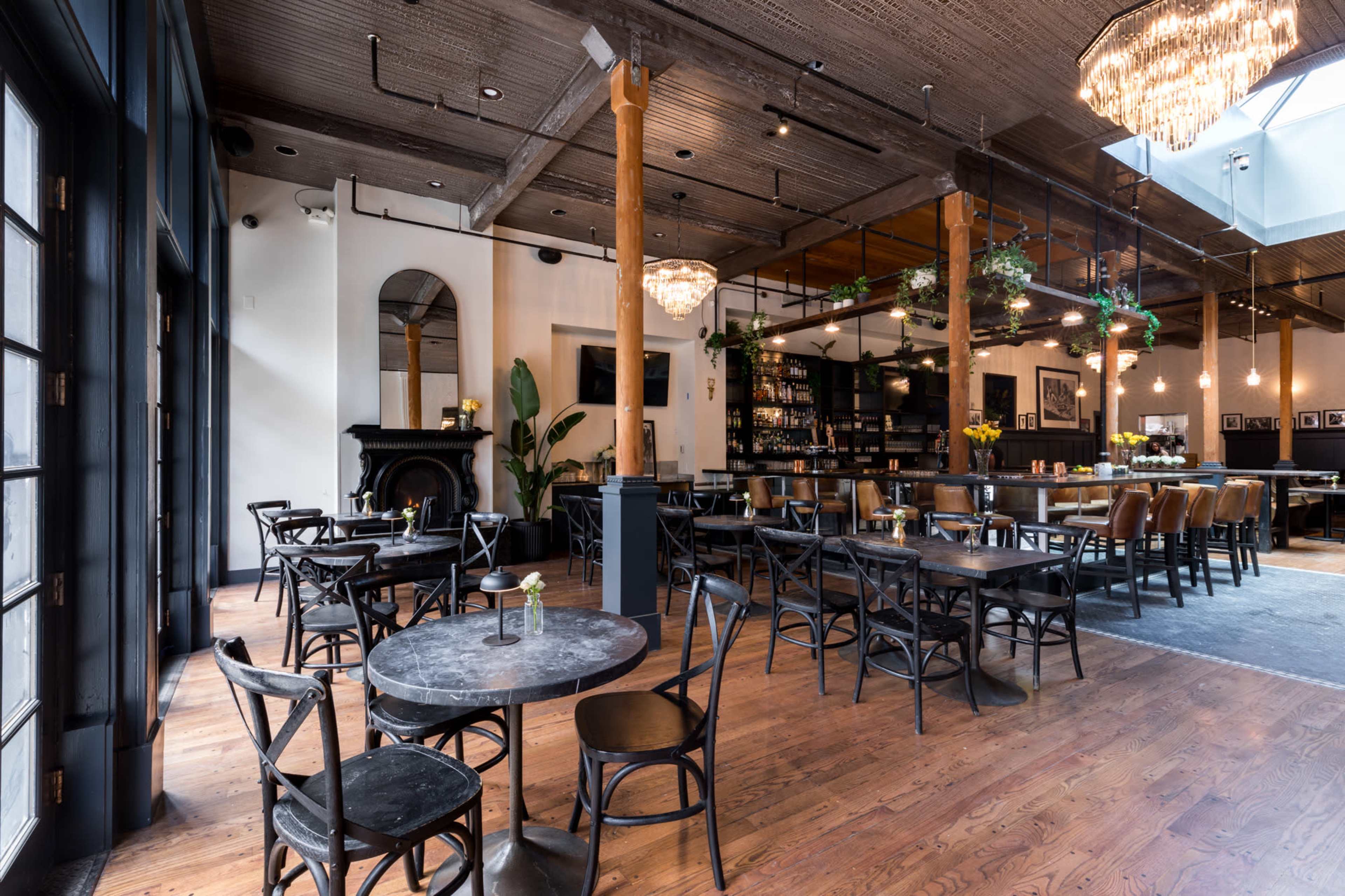 The image shows a spacious restaurant interior with wooden flooring, round tables, and a bar area, featuring large windows and a chandelier.