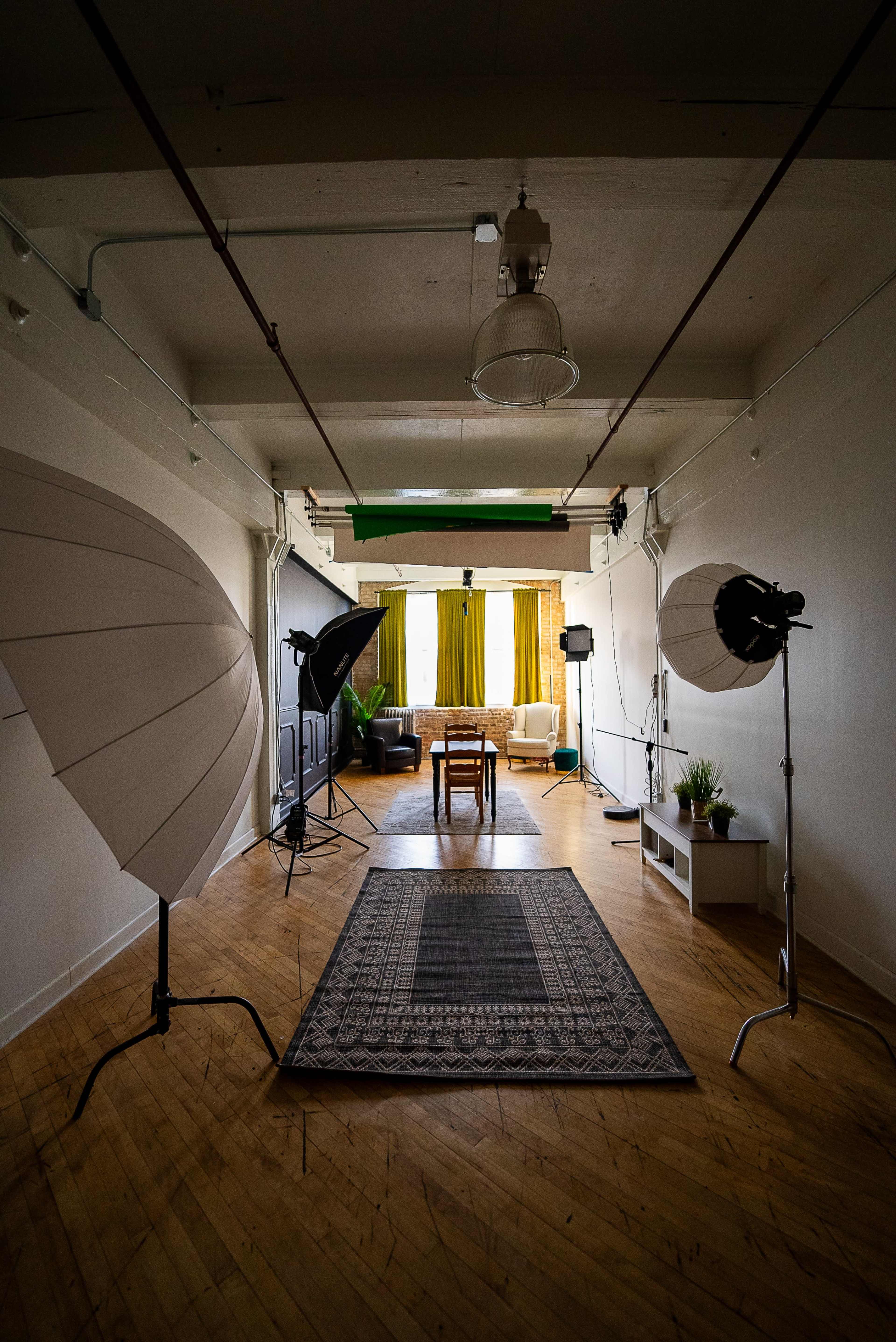 A spacious studio setup with softbox lights, a wooden table positioned on a patterned rug, and a seating area in the background framed by green curtains.