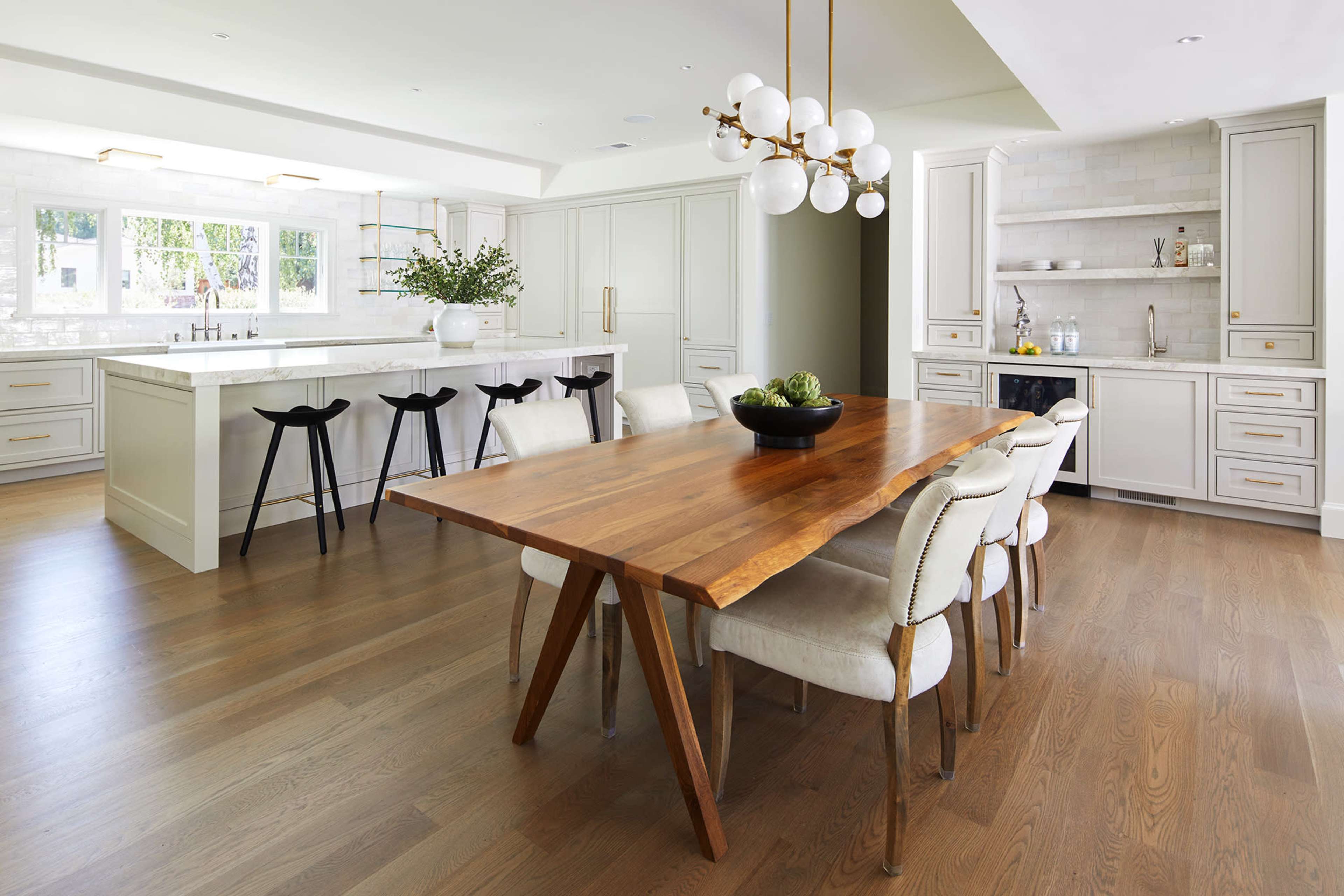 A modern kitchen and dining area featuring a long wooden table surrounded by white upholstered chairs, with a sleek kitchen island and cabinetry in the background.