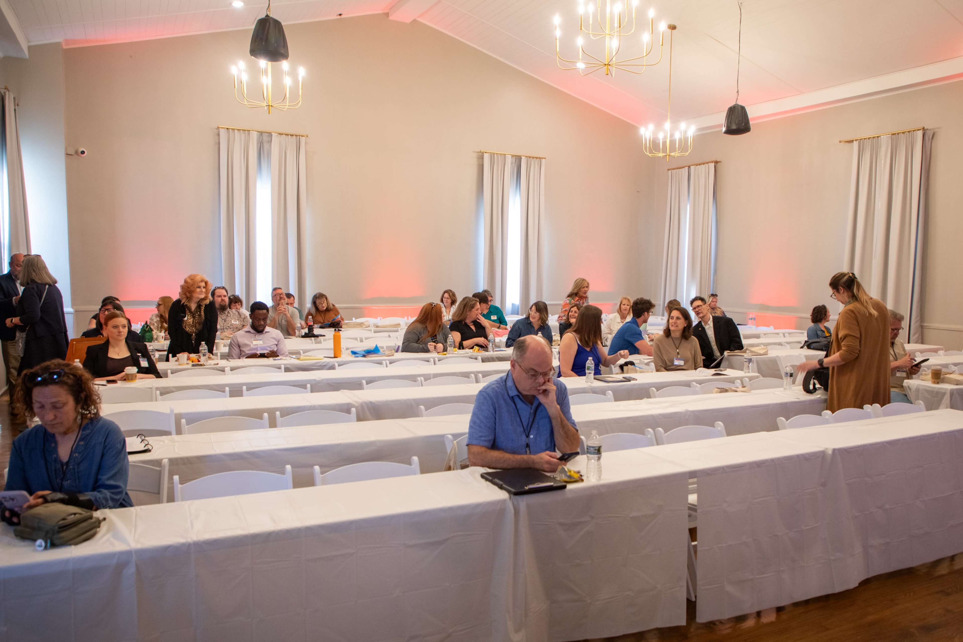A spacious conference room is set up with rows of tables covered in white tablecloths, and attendees are seated at some tables while others are empty.