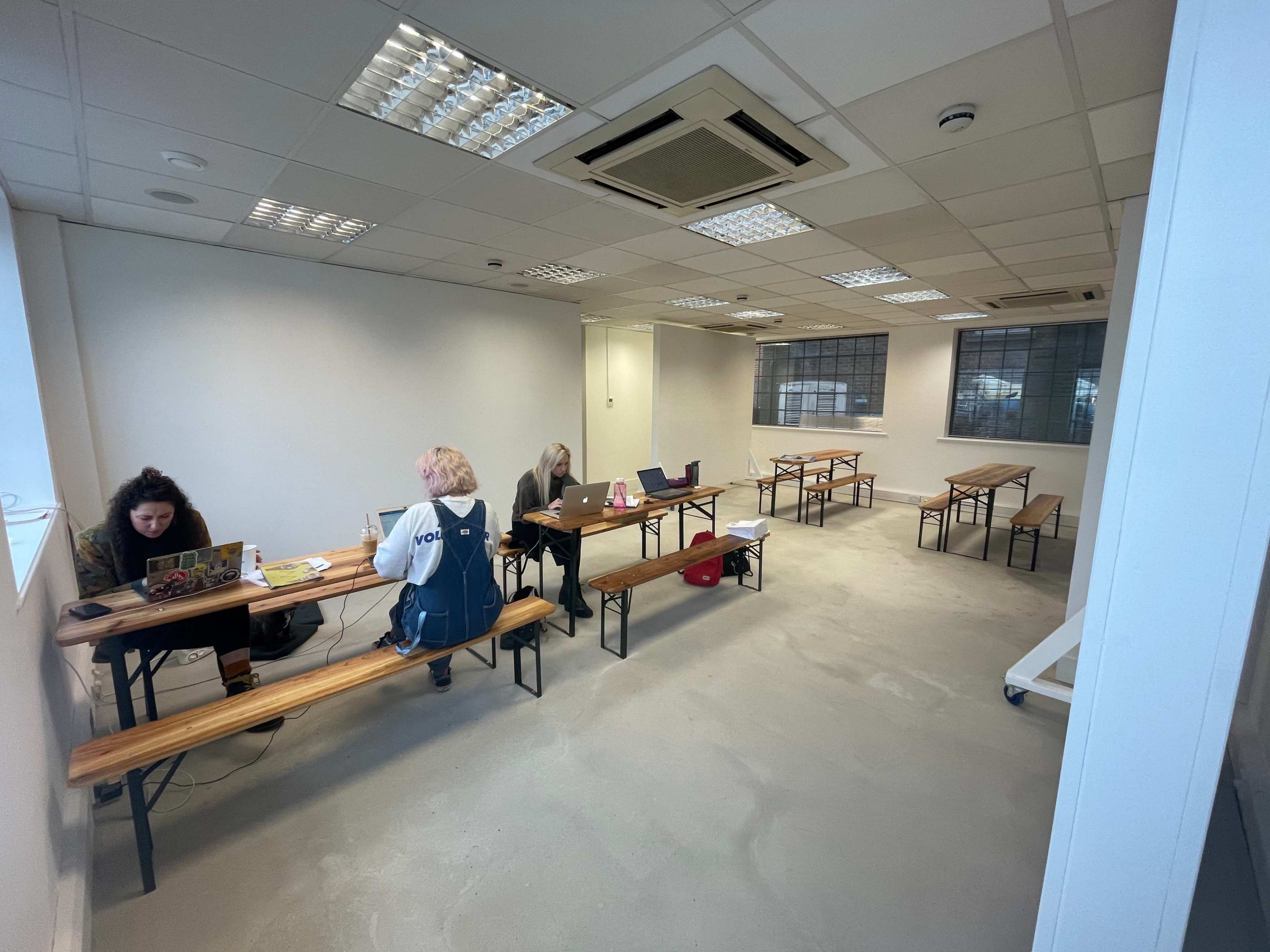 A sparsely furnished room with three individuals working on laptops at long wooden tables.