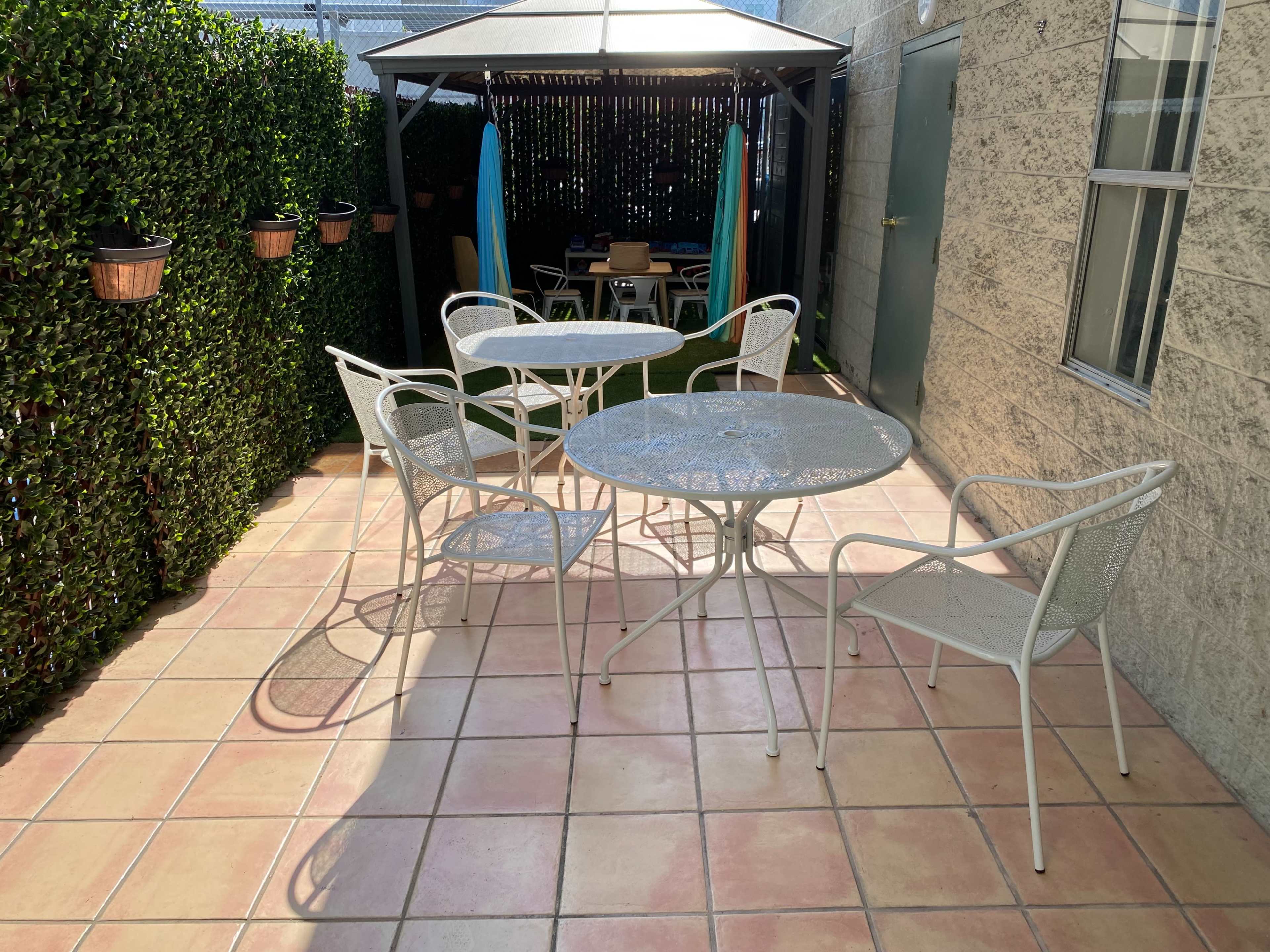 A patio area features a glass-top table surrounded by four white metal chairs, with a gazebo in the background and potted plants along the walls.