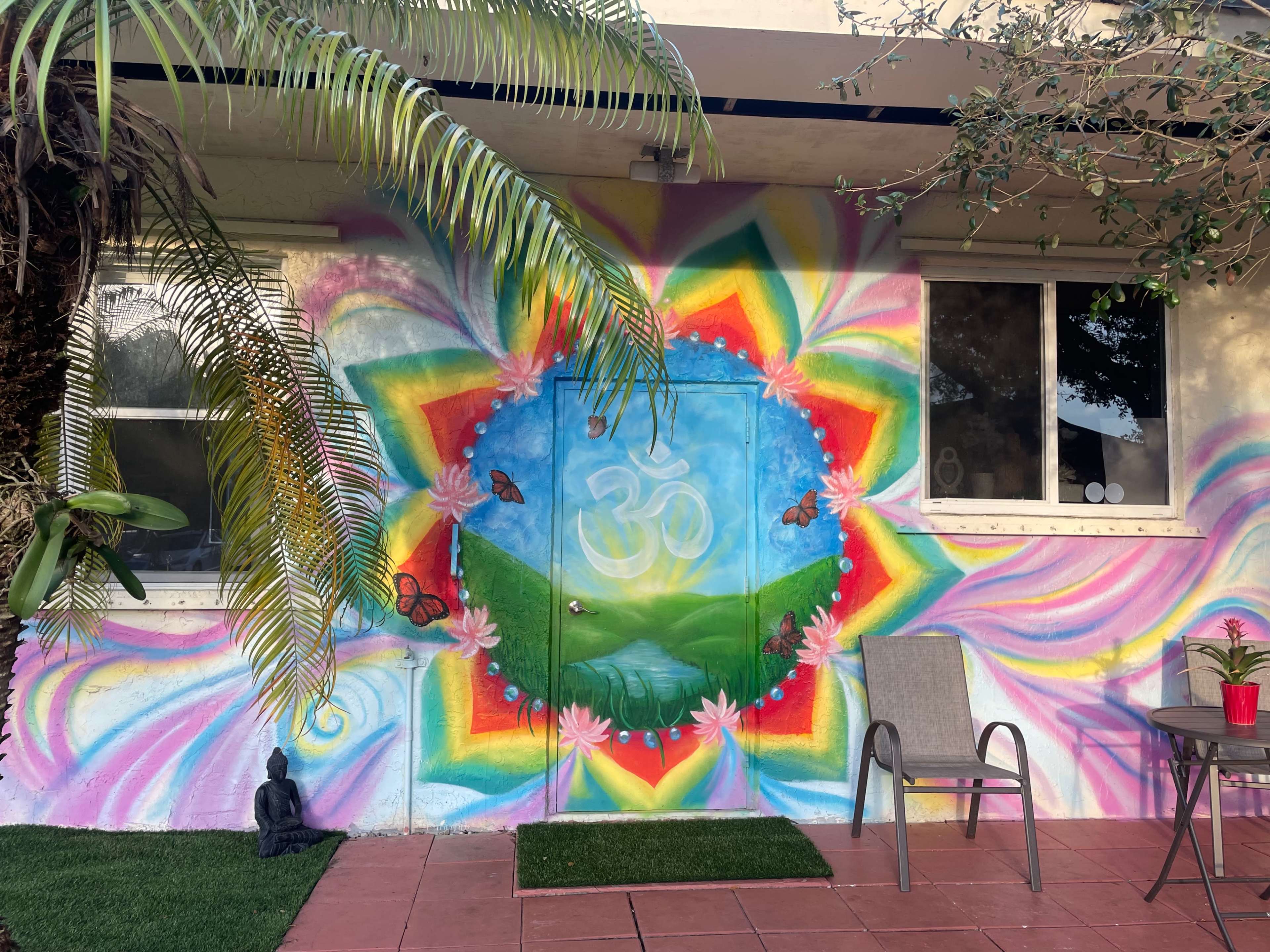 The image shows a colorful mural featuring a large "Om" symbol surrounded by butterflies and lotus flowers, with a palm tree to the left and a small seating area with a plant in front of it.
