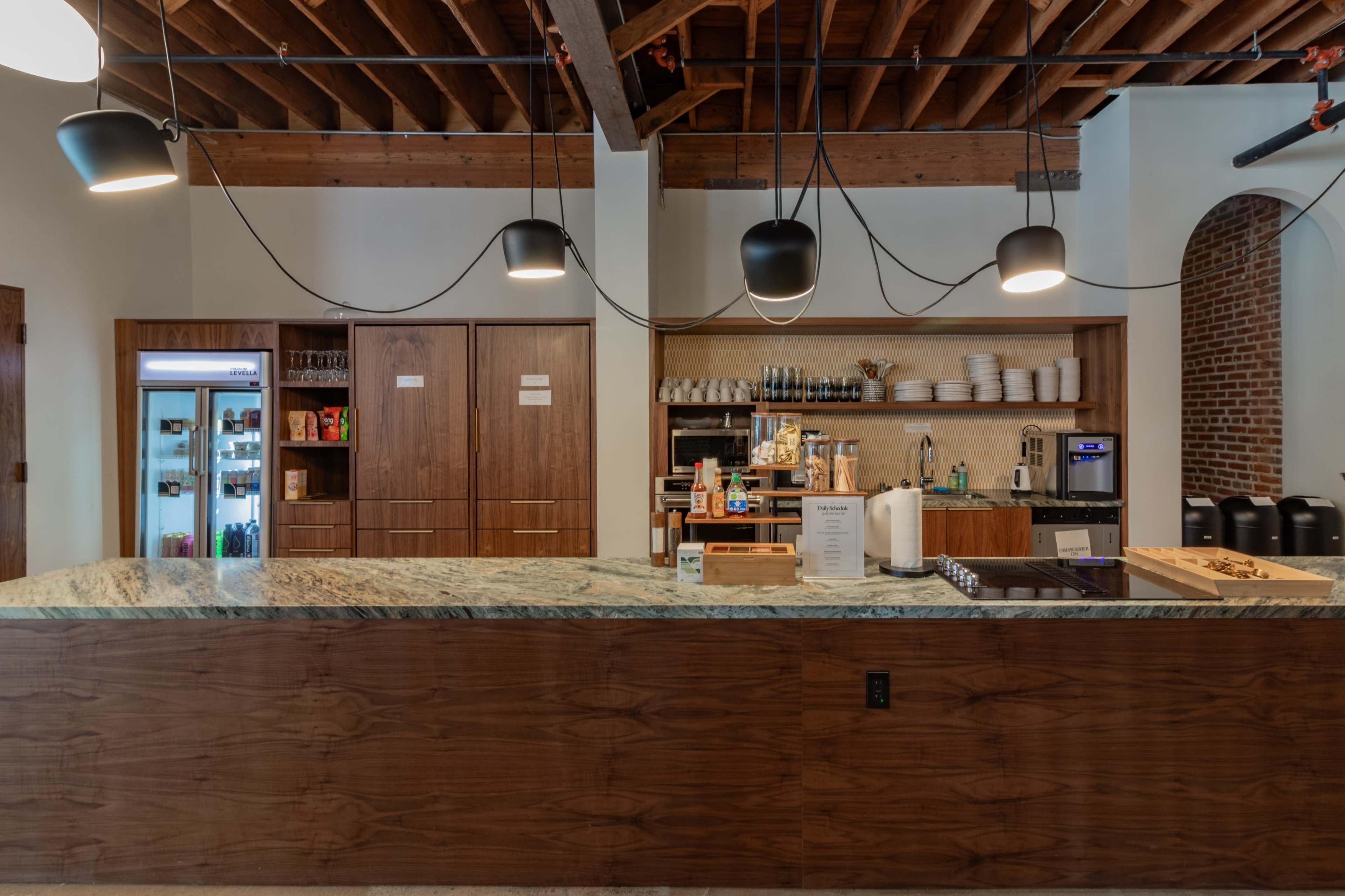 A modern, minimalist cafe interior featuring a wooden counter with a granite top, overhead pendant lights, and shelves stocked with dishes and beverages.