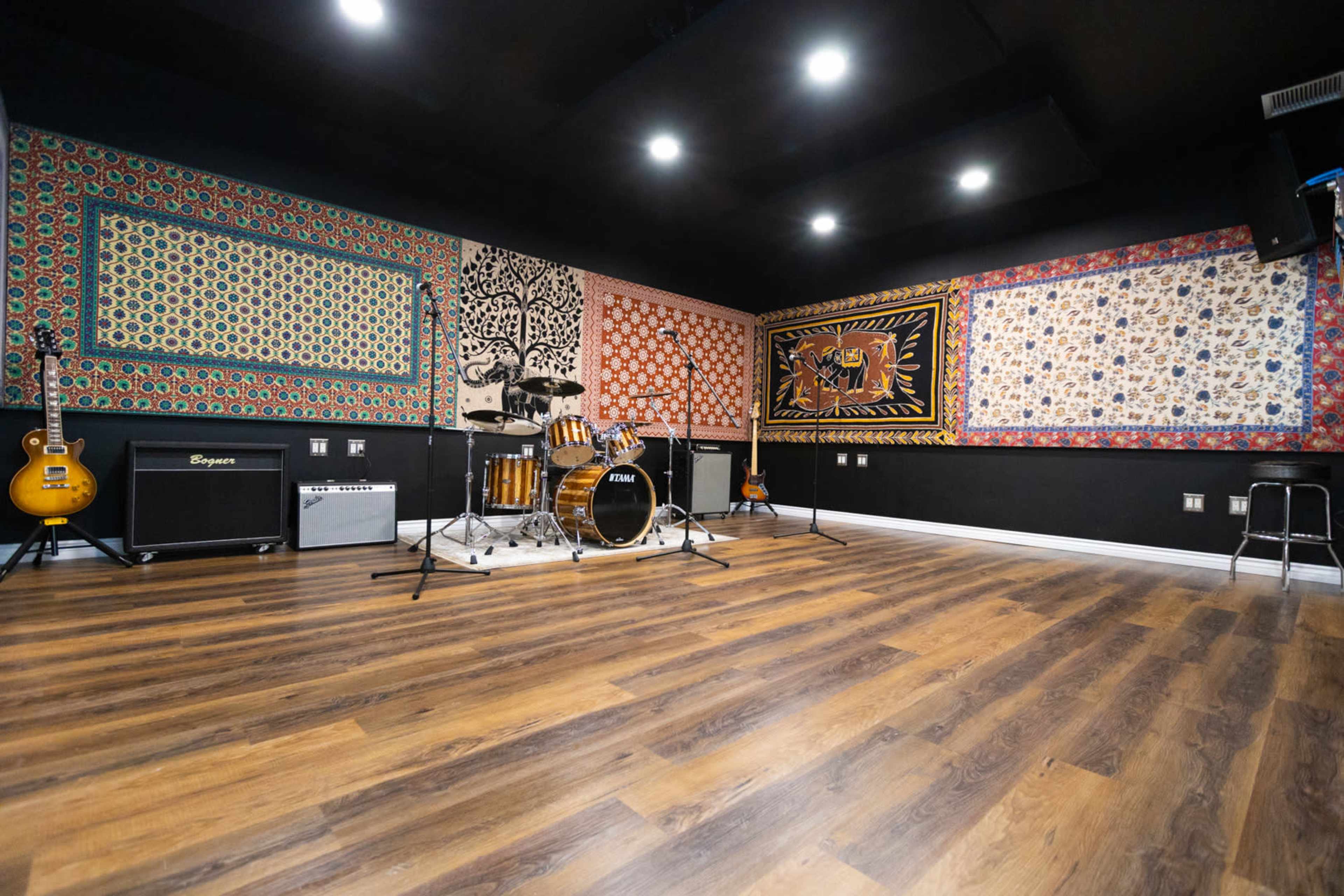 The image shows a music rehearsal space featuring a drum set, guitars, amplifiers, and colorful wall tapestries.