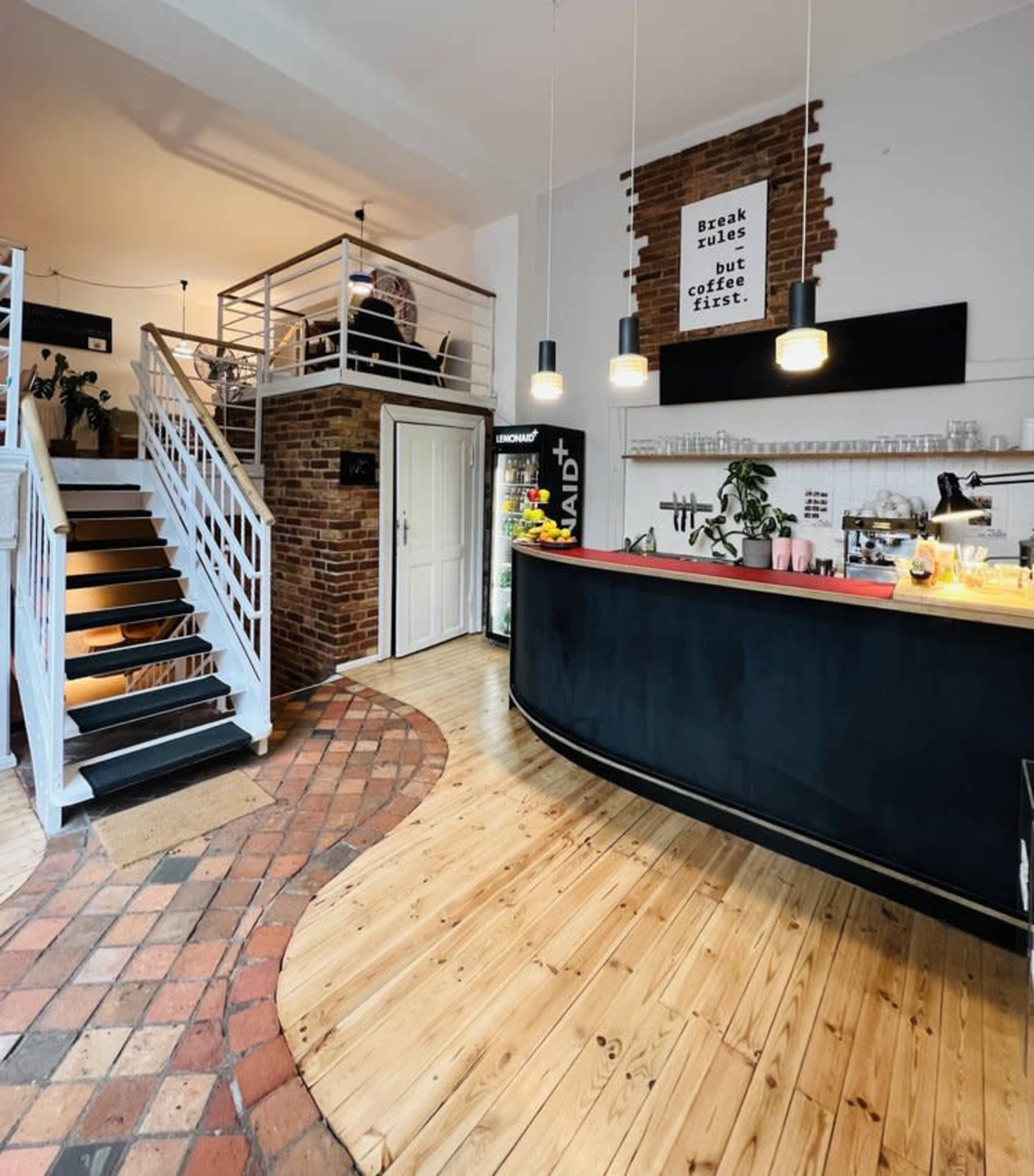 The image shows a spacious café interior featuring a wooden floor, a curved counter, and a staircase leading to a second level with a seating area.
