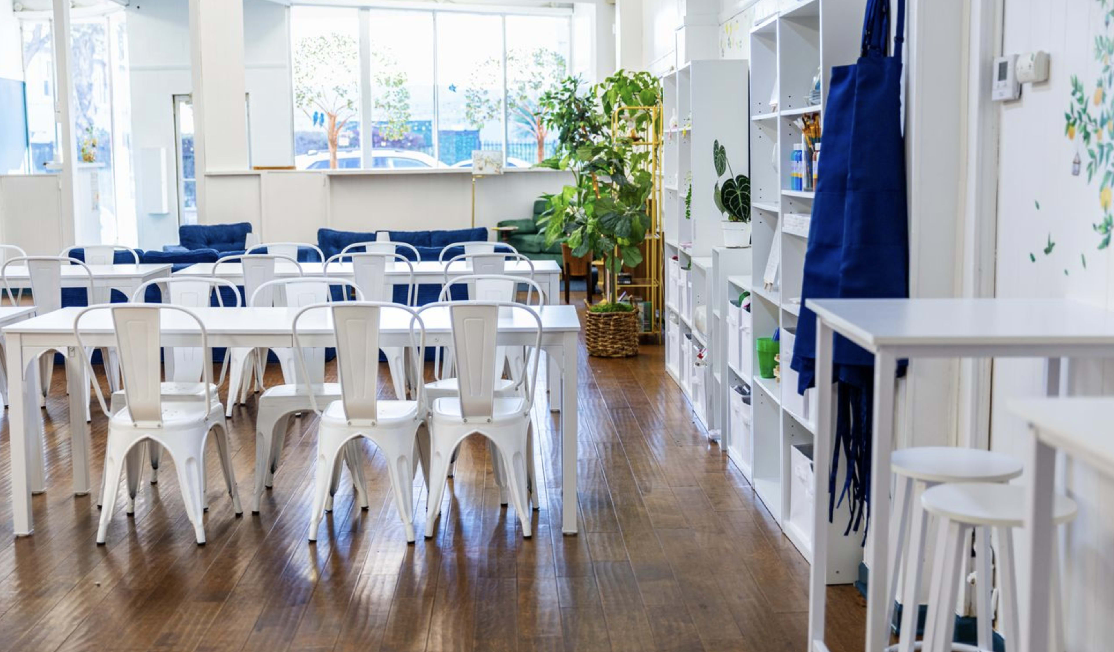 The image shows a bright, open interior space with white tables and chairs, shelves lined with plants and decorative items, and large windows allowing natural light to enter.
