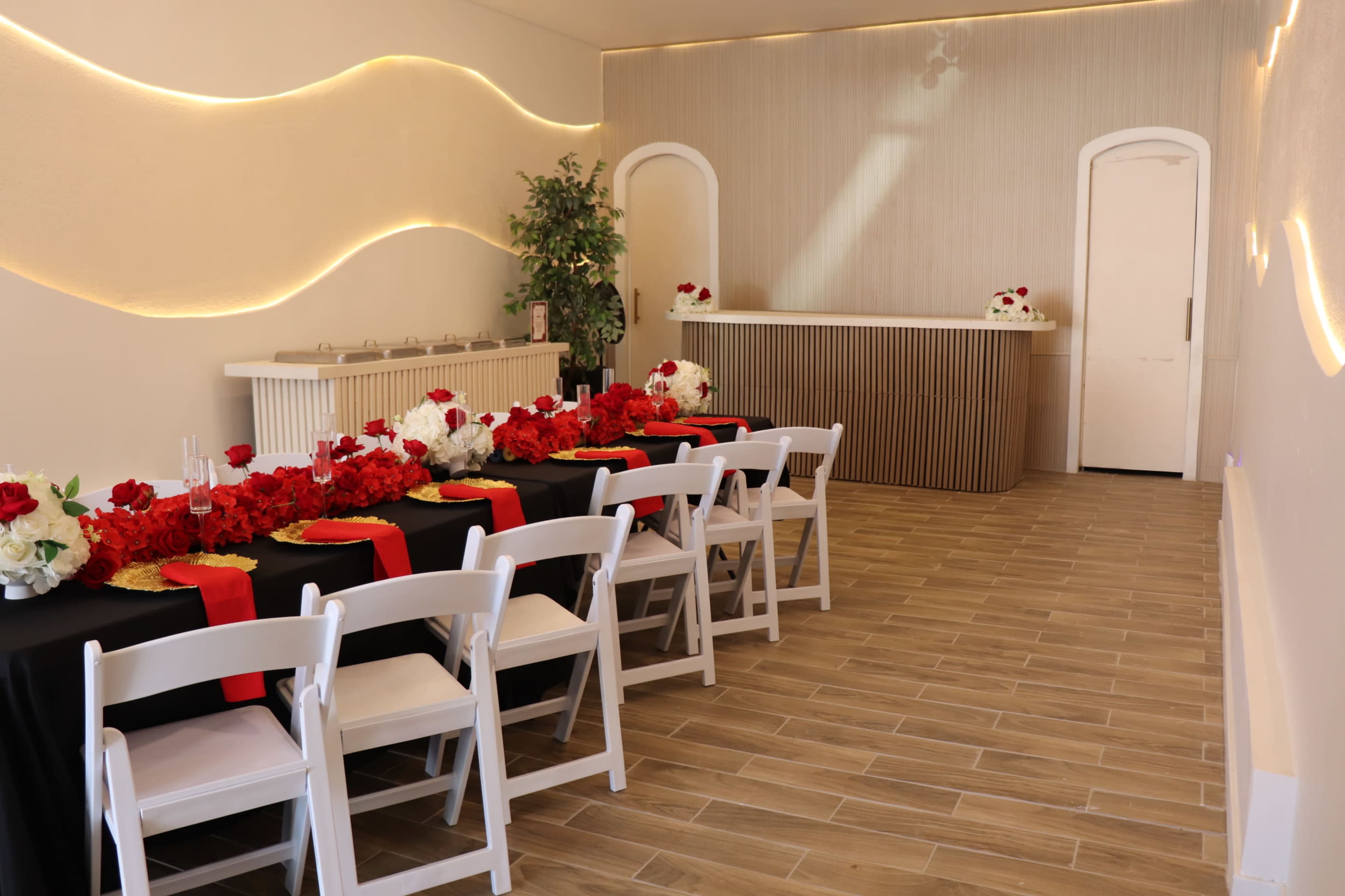 A dining area is set with a long table adorned with red roses and black tablecloths, surrounded by white chairs, and a bar area featuring a light wood design.