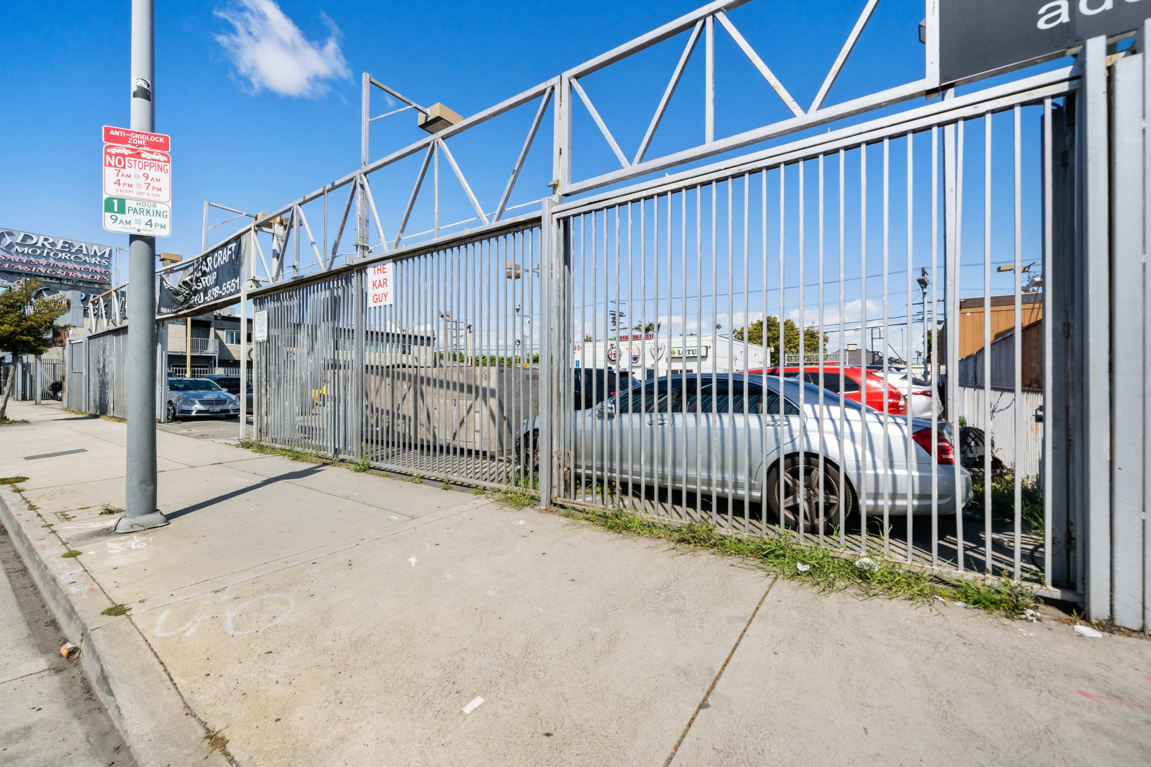 A silver car is parked behind a metal fence next to a sidewalk, with a clear blue sky overhead.