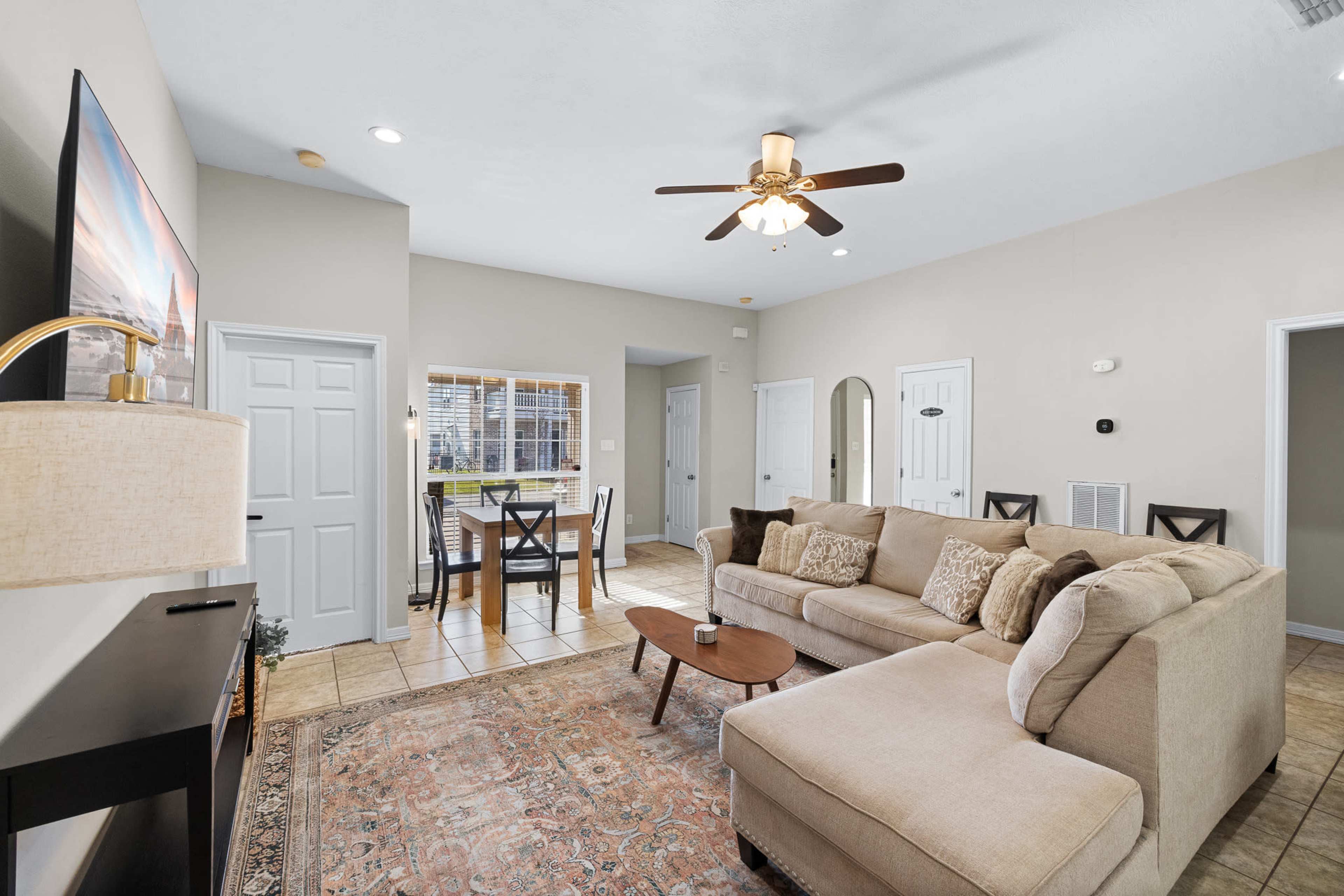 The living room features a beige sectional sofa, a wooden coffee table, and a ceiling fan, with a dining area visible in the background.