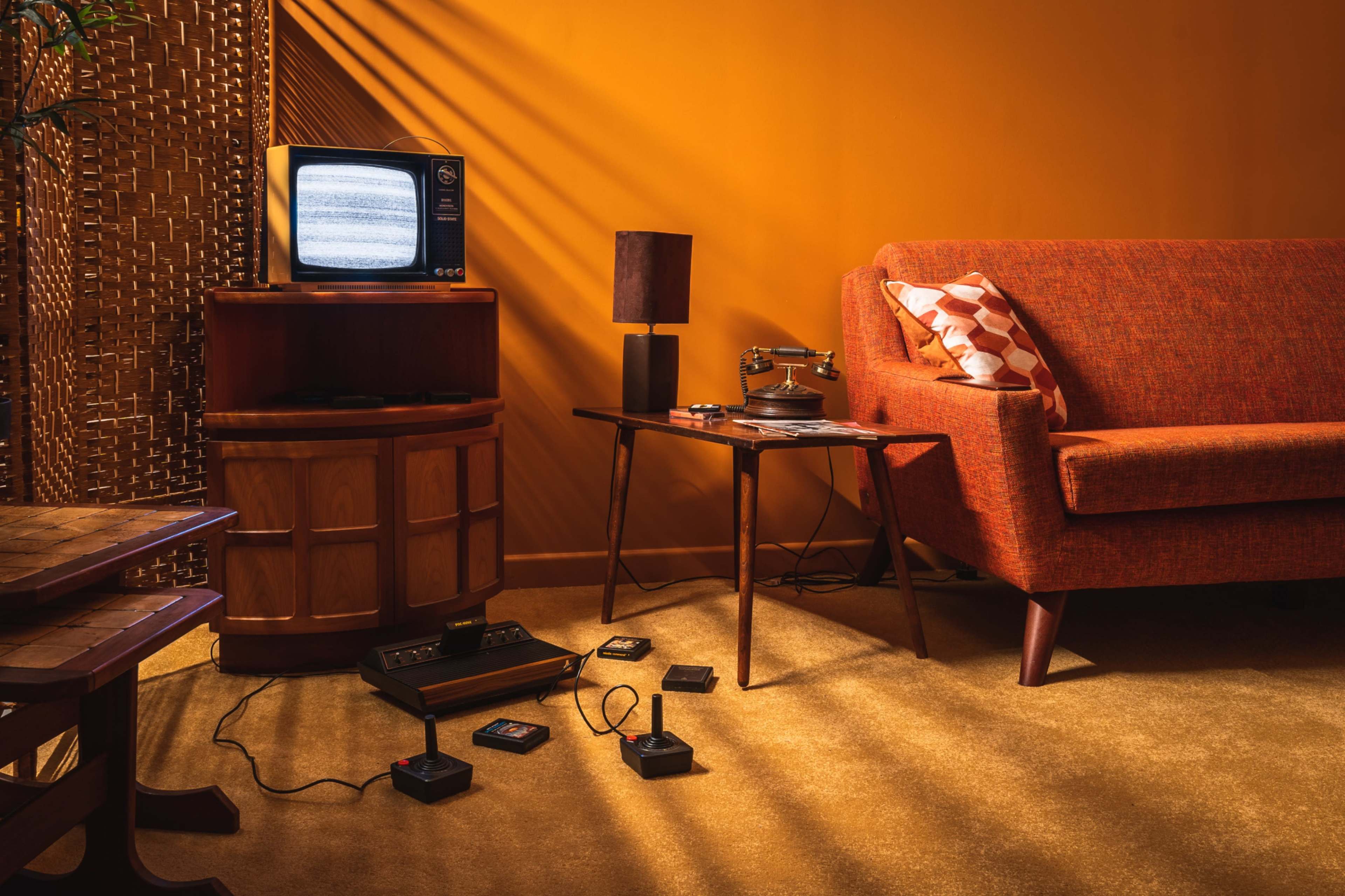 The scene features a vintage living room with an orange wall, a tube television displaying static, an orange couch, a lamp, and an Atari gaming console connected to several controllers on the carpeted floor.