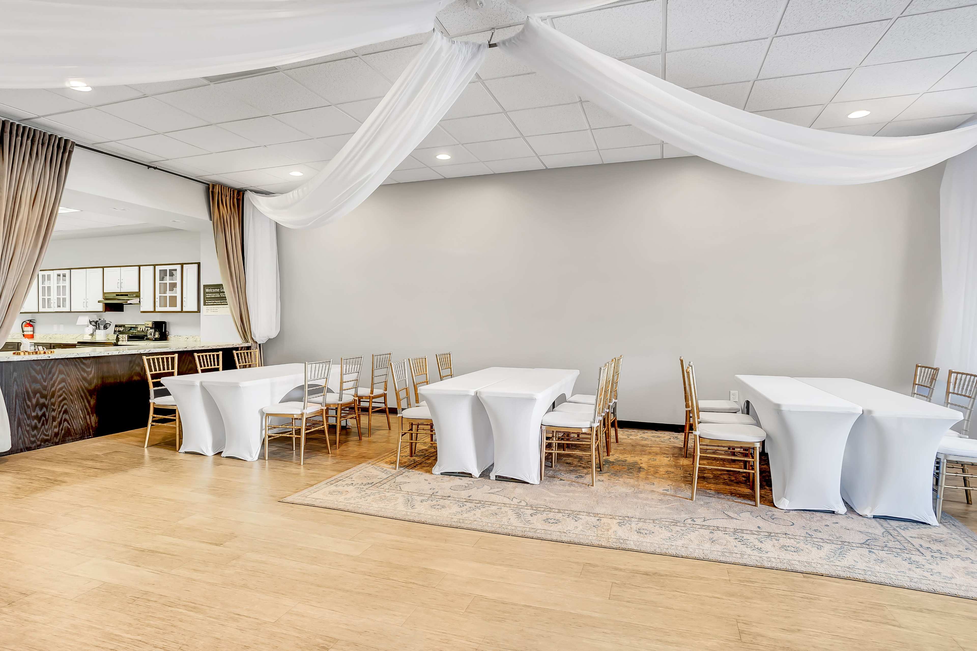 A spacious event room with several white-covered tables arranged neatly, draped fabric on the ceiling, and a bar area in the background.