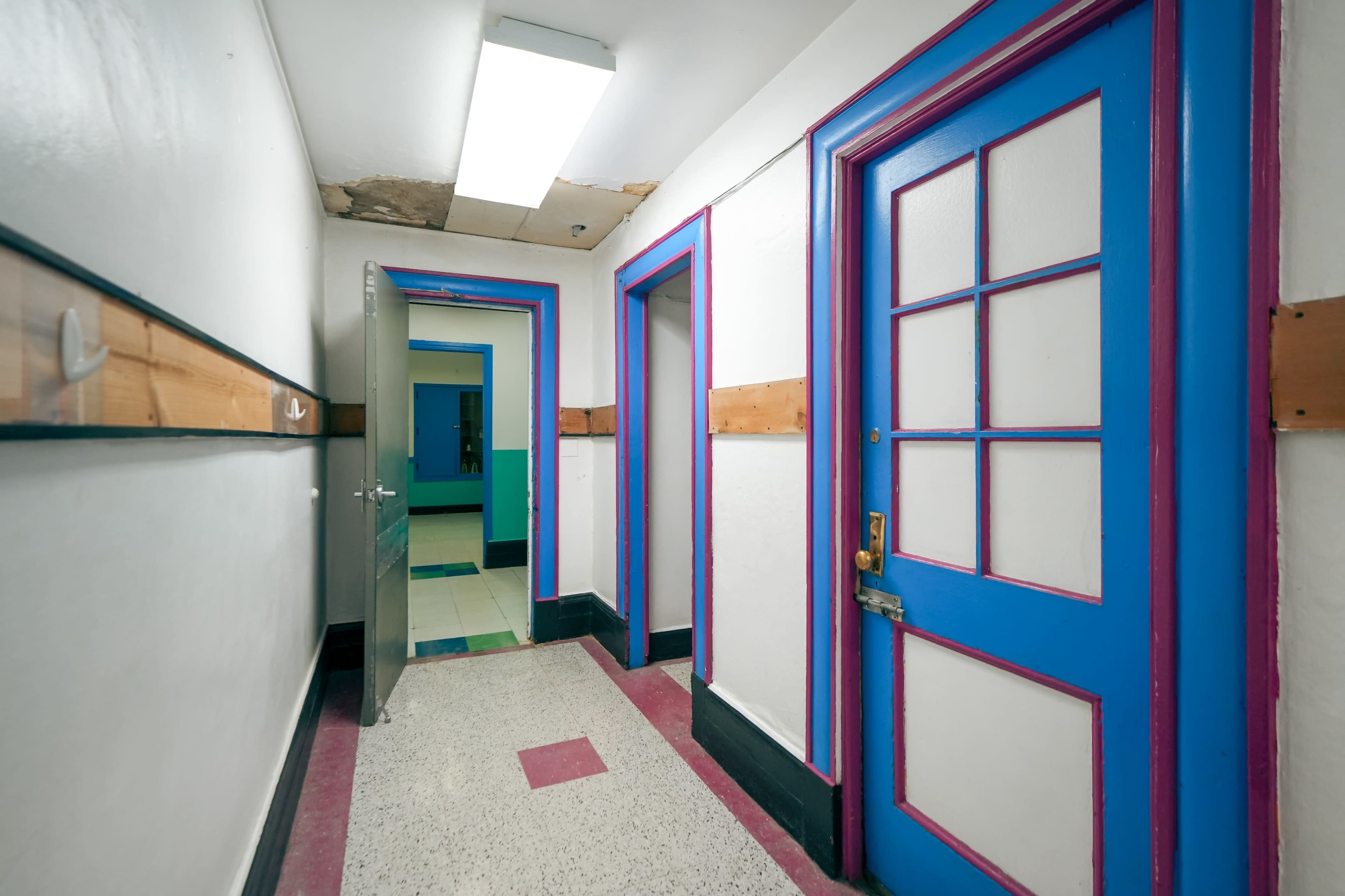 The image shows a colorful hallway with blue doors and light-colored walls, featuring a patterned floor and a visible open door at the end.