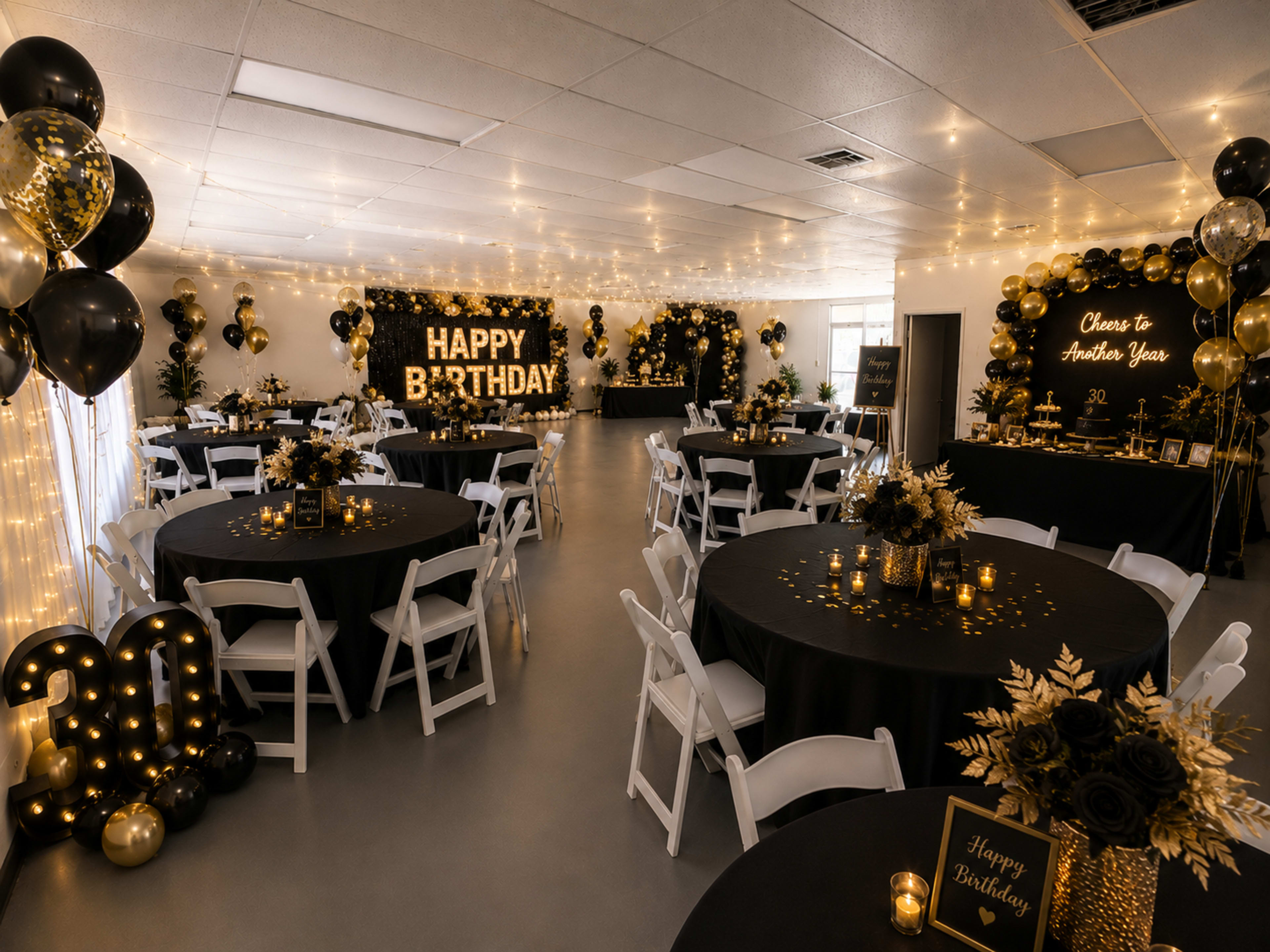 The image shows a spacious event room decorated for a birthday celebration, featuring black and gold balloons, tables with black tablecloths, and a large "HAPPY BIRTHDAY" sign illuminated on the wall.