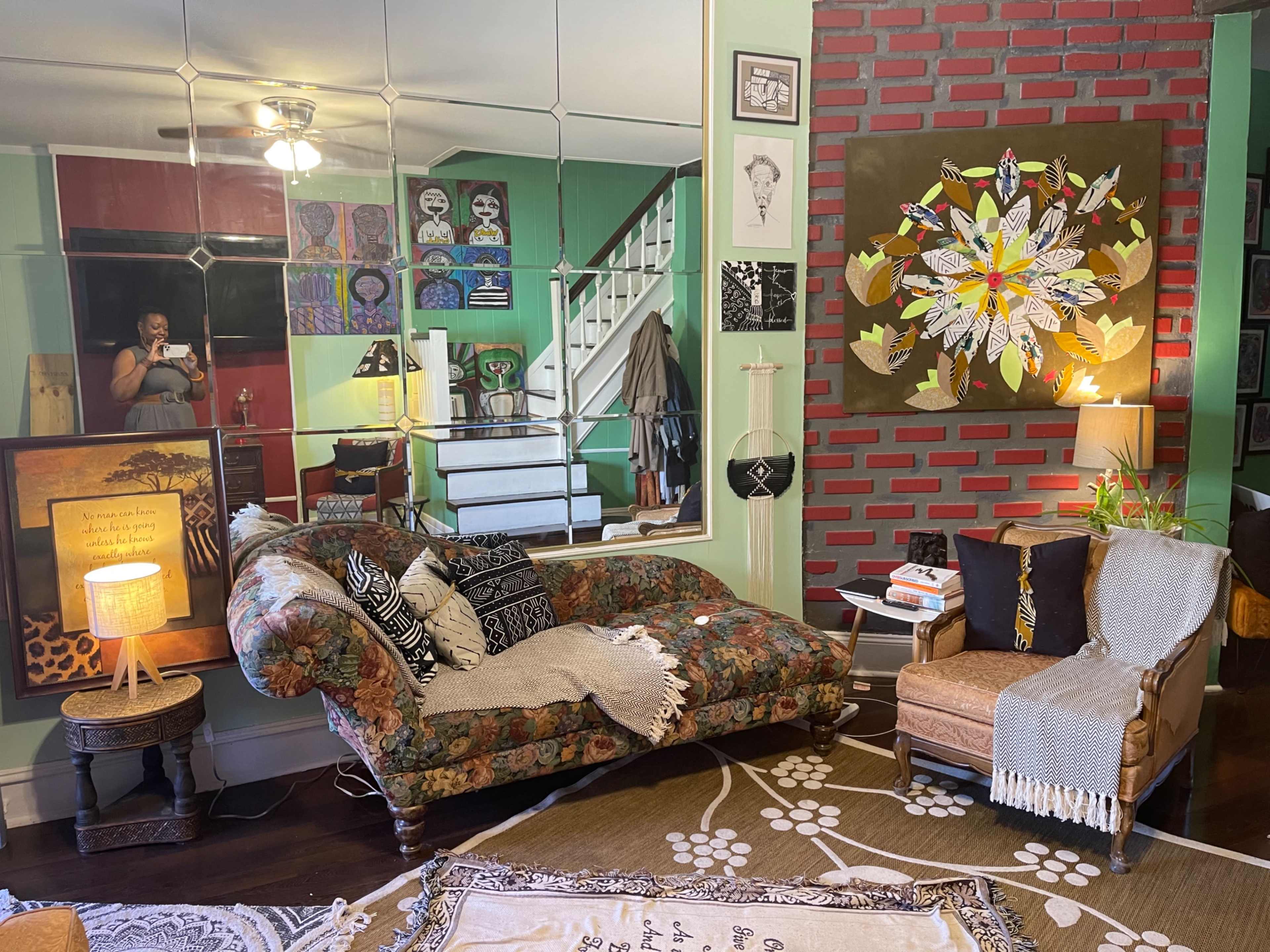The image depicts a living room with a mirrored wall, a floral couch, a wooden chair, and colorful artwork against a brick accent wall.