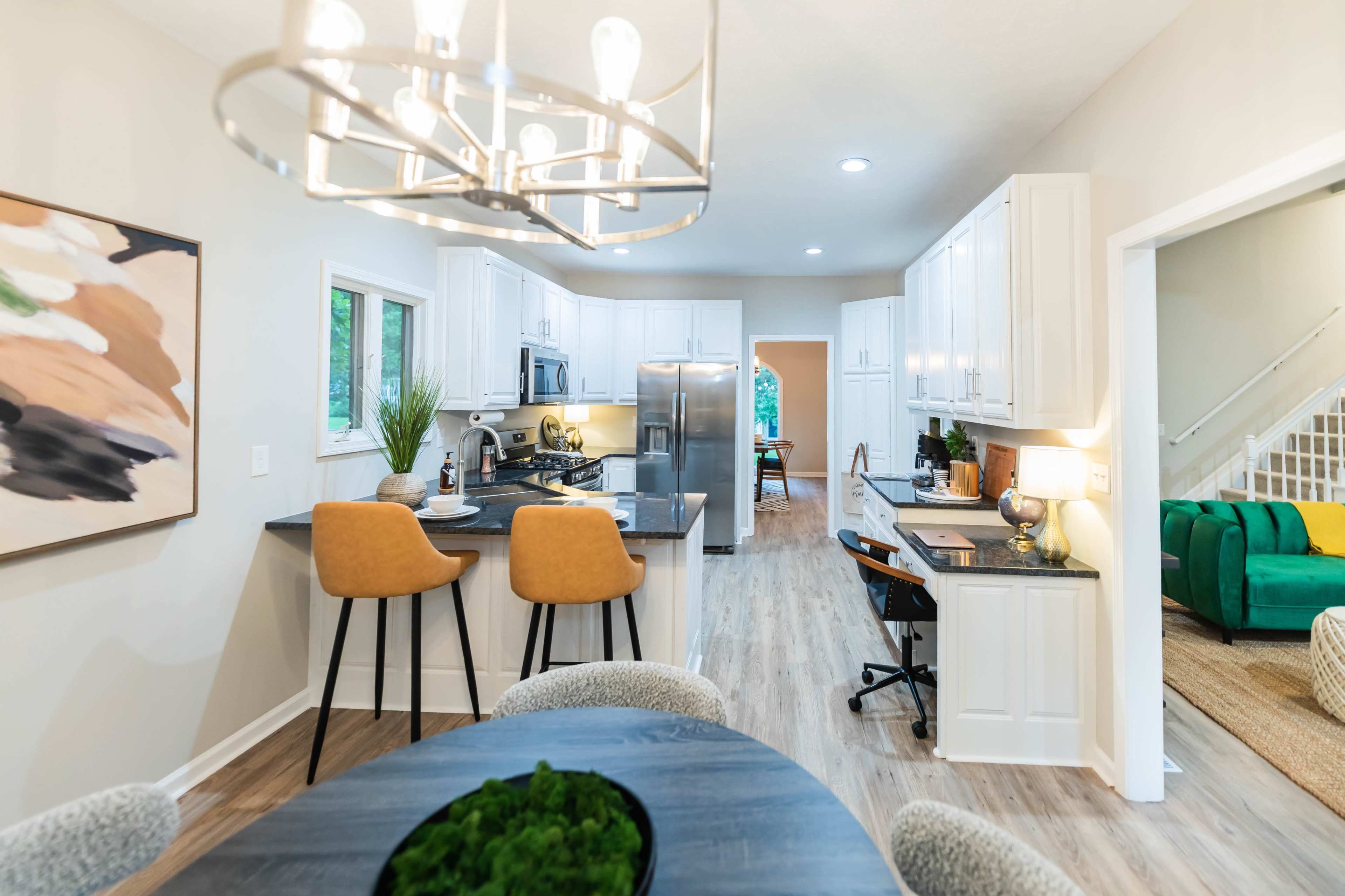A modern kitchen and dining area featuring white cabinets, a central island with two chairs, and a visible workspace leading to another room.