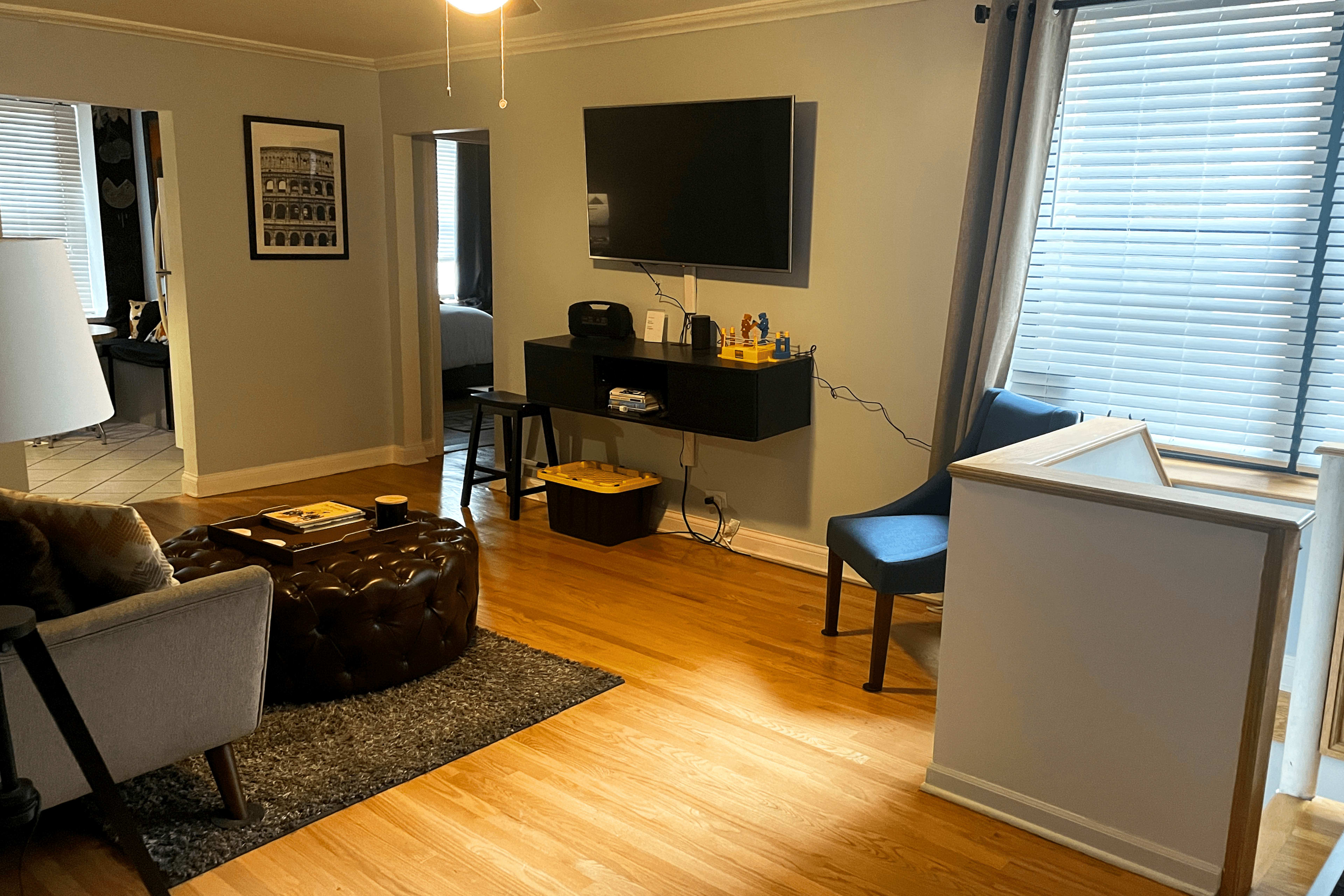 A living room with hardwood floors, a television mounted on the wall, a dark ottoman, a blue chair, and a window with blinds.