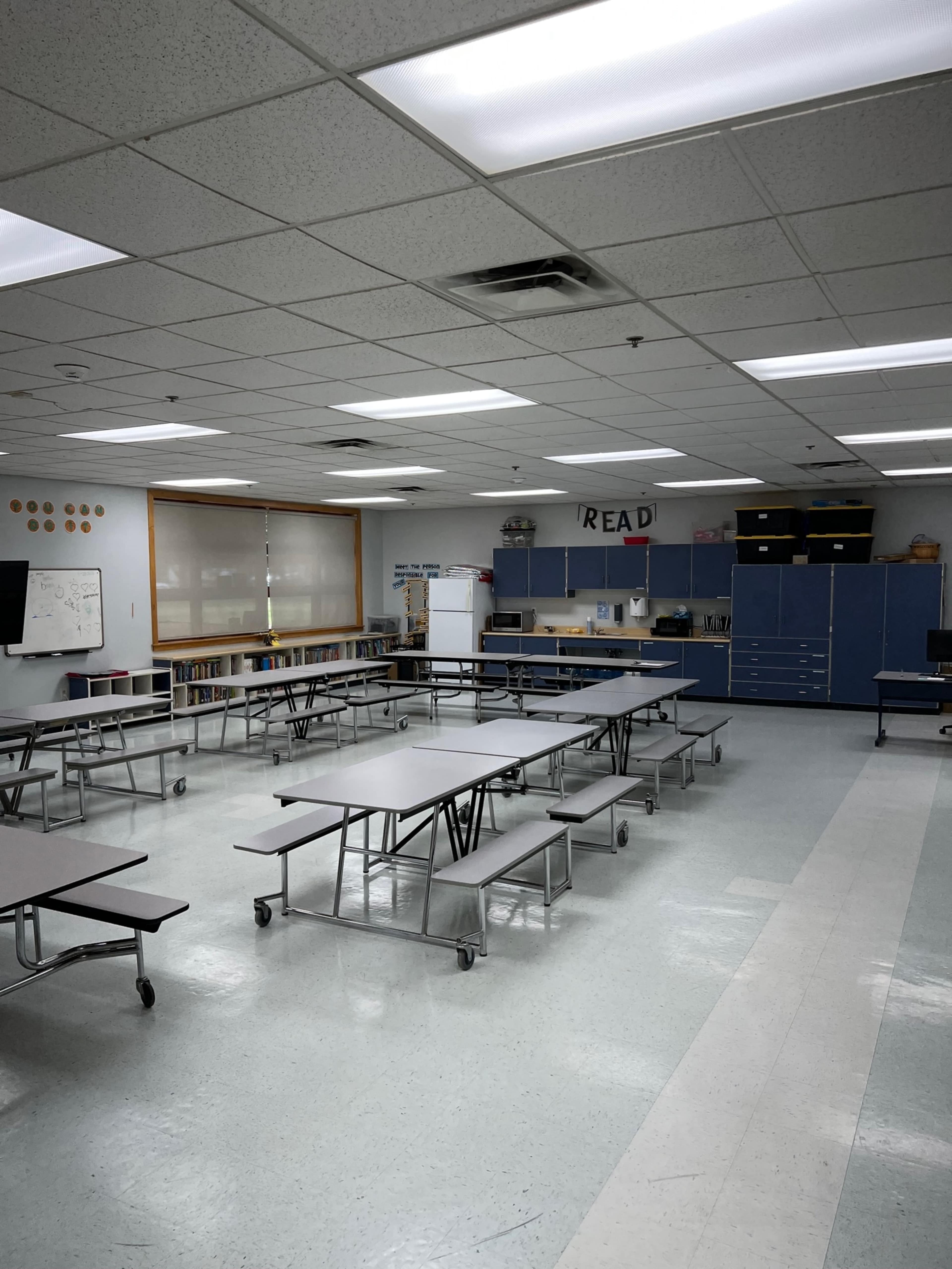 An empty classroom with rows of tables arranged in the center, blue cabinets lining the walls, and shelves filled with books.