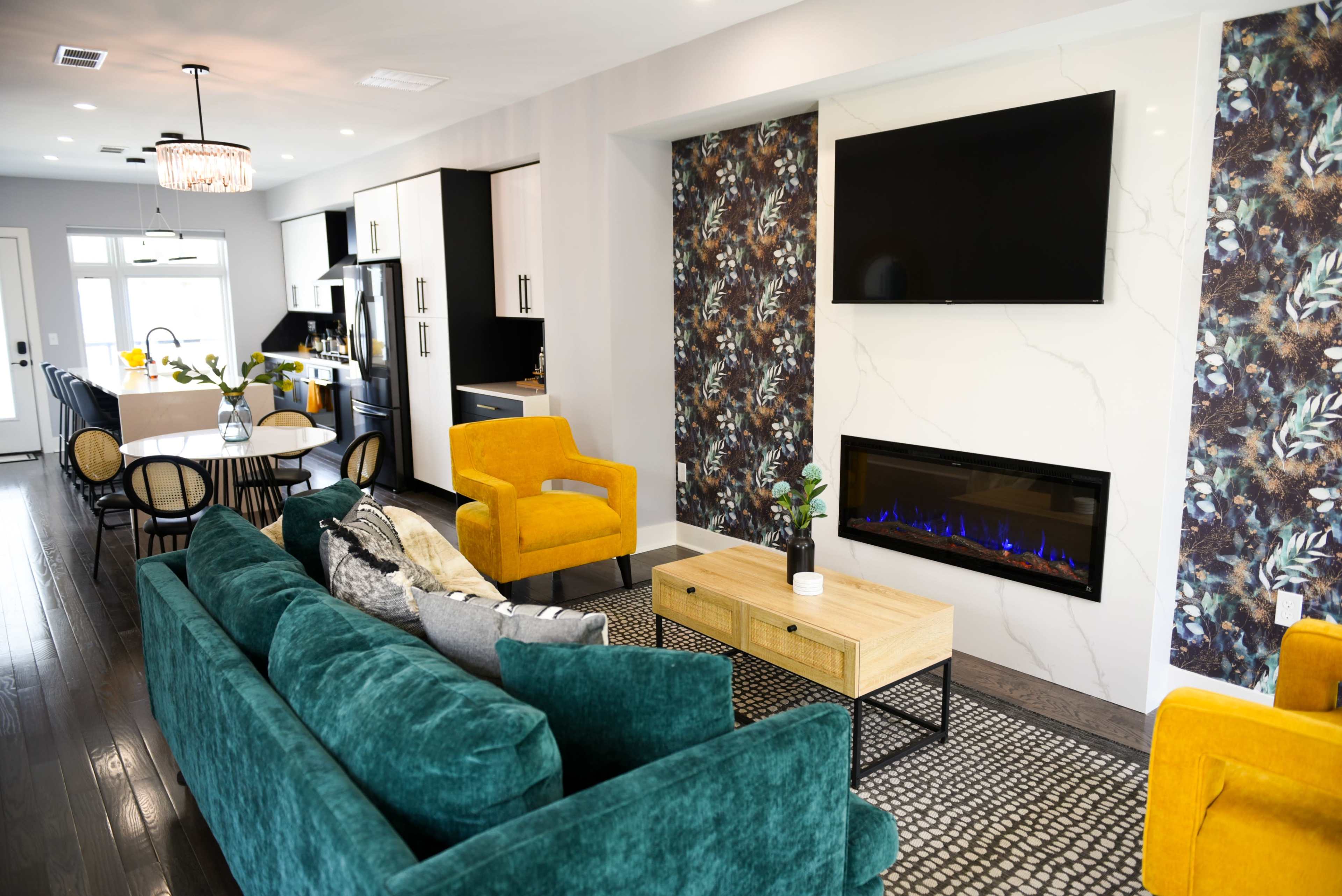 A modern living room featuring a teal sofa, yellow armchairs, a wooden coffee table, and a wall-mounted TV above a fireplace.