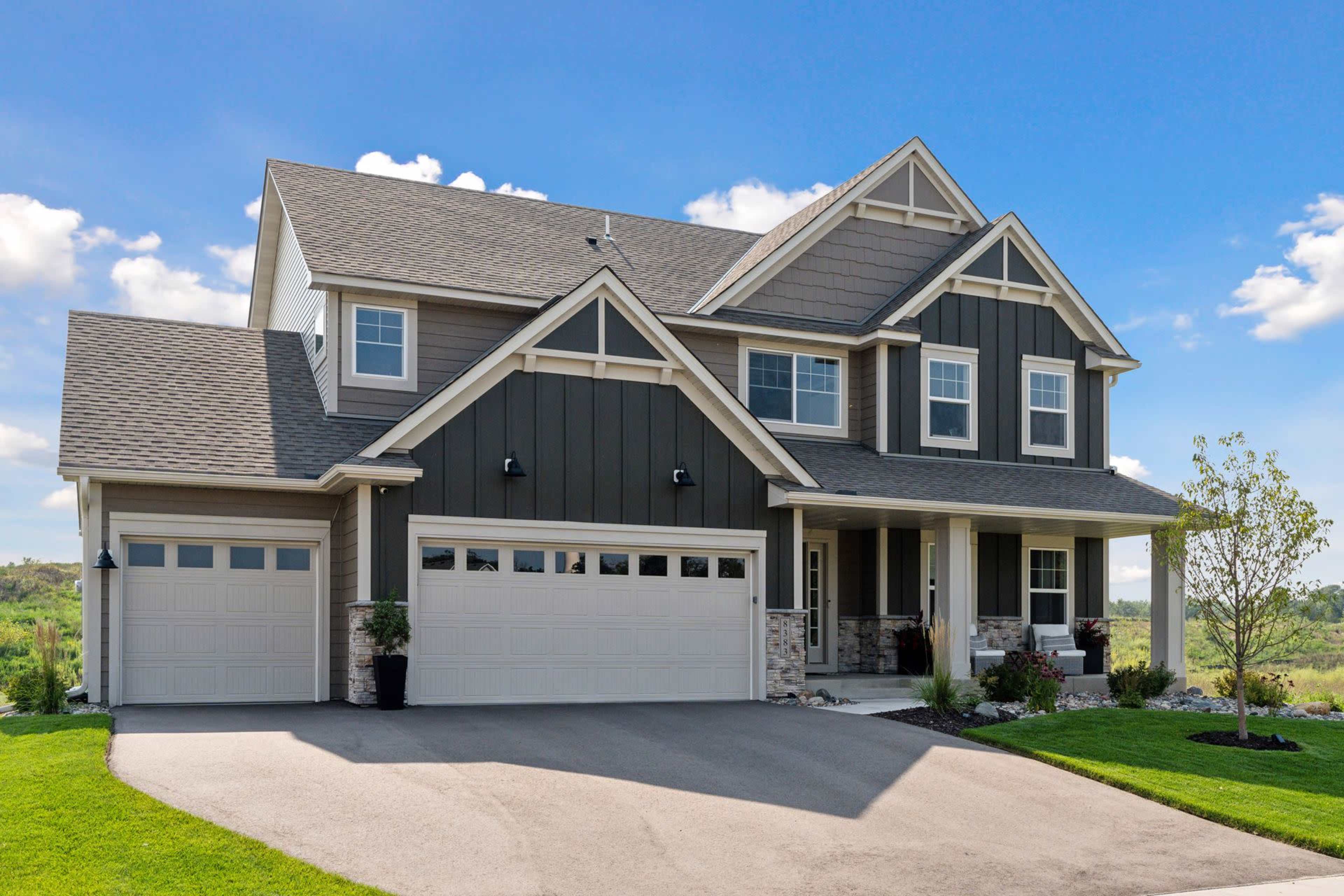 The image shows a two-story house with a dark exterior, two garages, and a landscaped front yard under a clear blue sky.